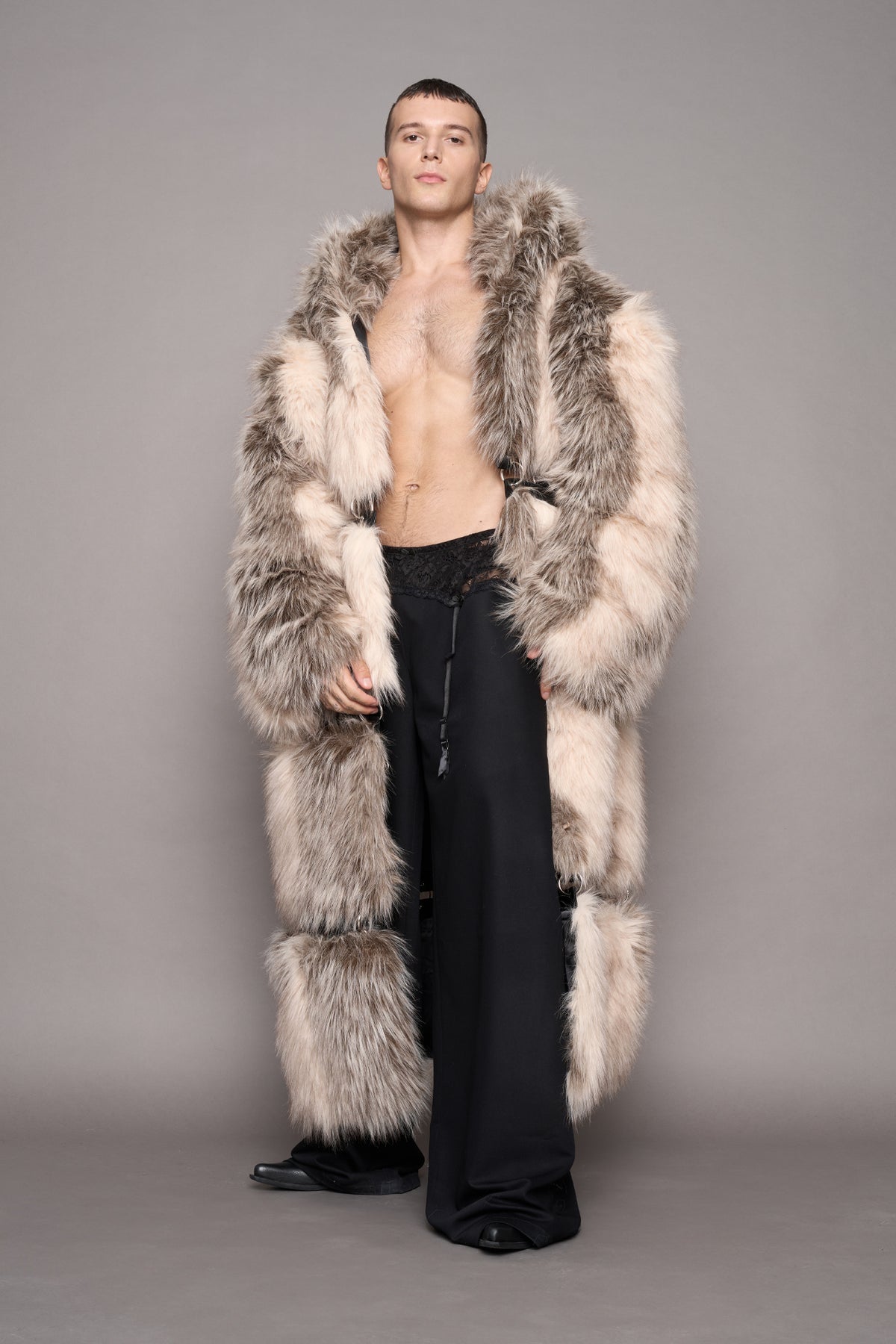 A male model stands against a plain gray studio background wearing a very large shaggy faux-fur coat in mixed beige, cream, and gray tones over black wide-leg pants and black shoes.