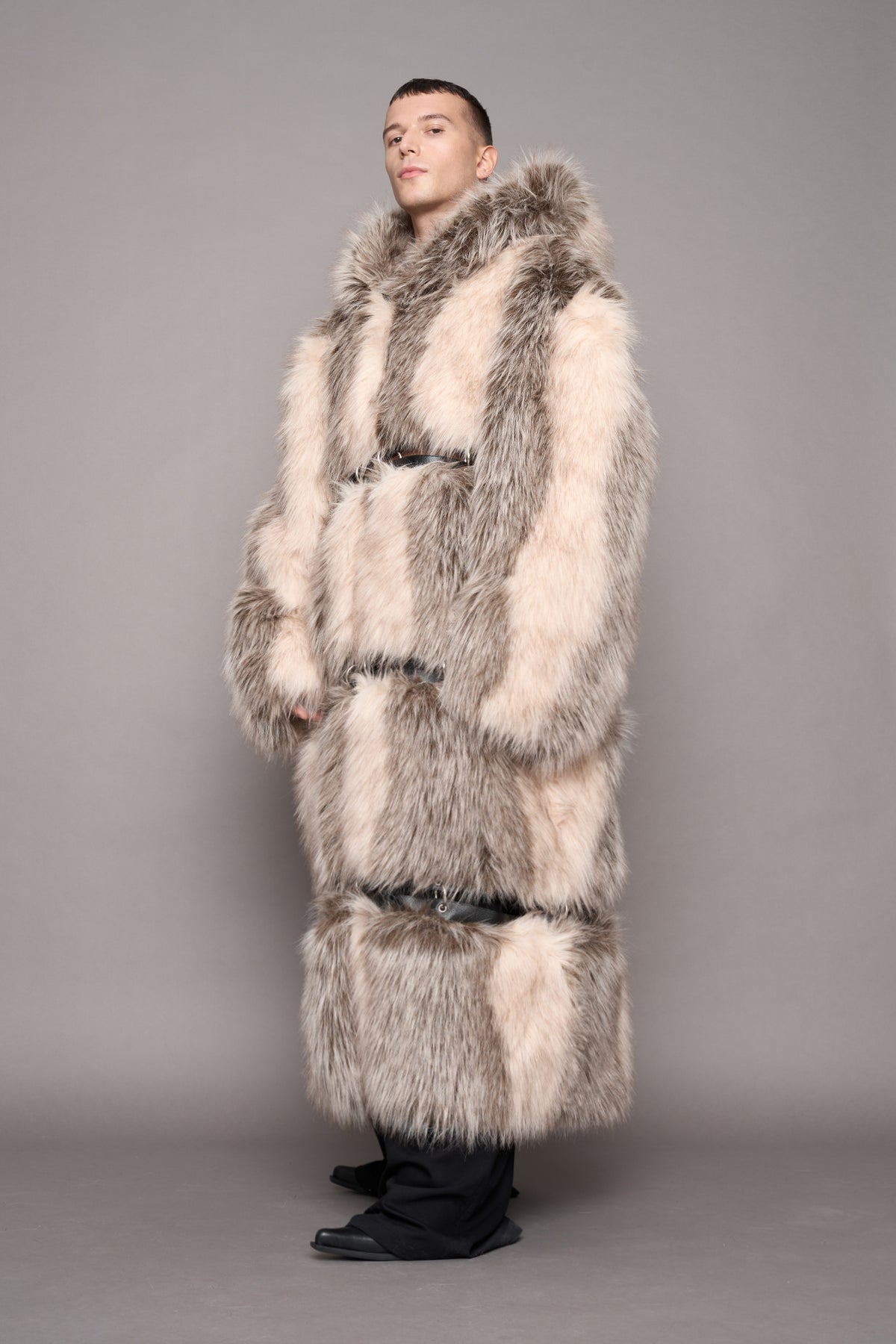 A model wearing a long, chunky faux fur coat in shades of beige, gray, and brown, styled with black trousers and shoes against a solid gray studio background.