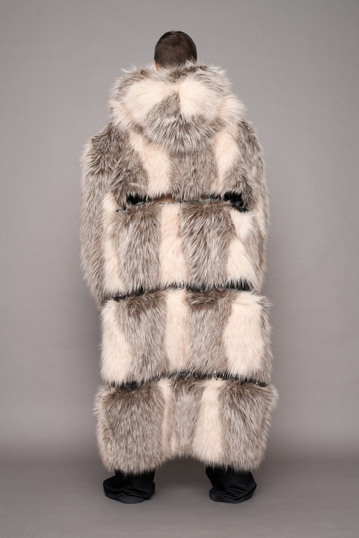 A person viewed from behind wearing a long, oversized faux fur coat in light gray, cream, and white tones, standing against a plain gray studio background.