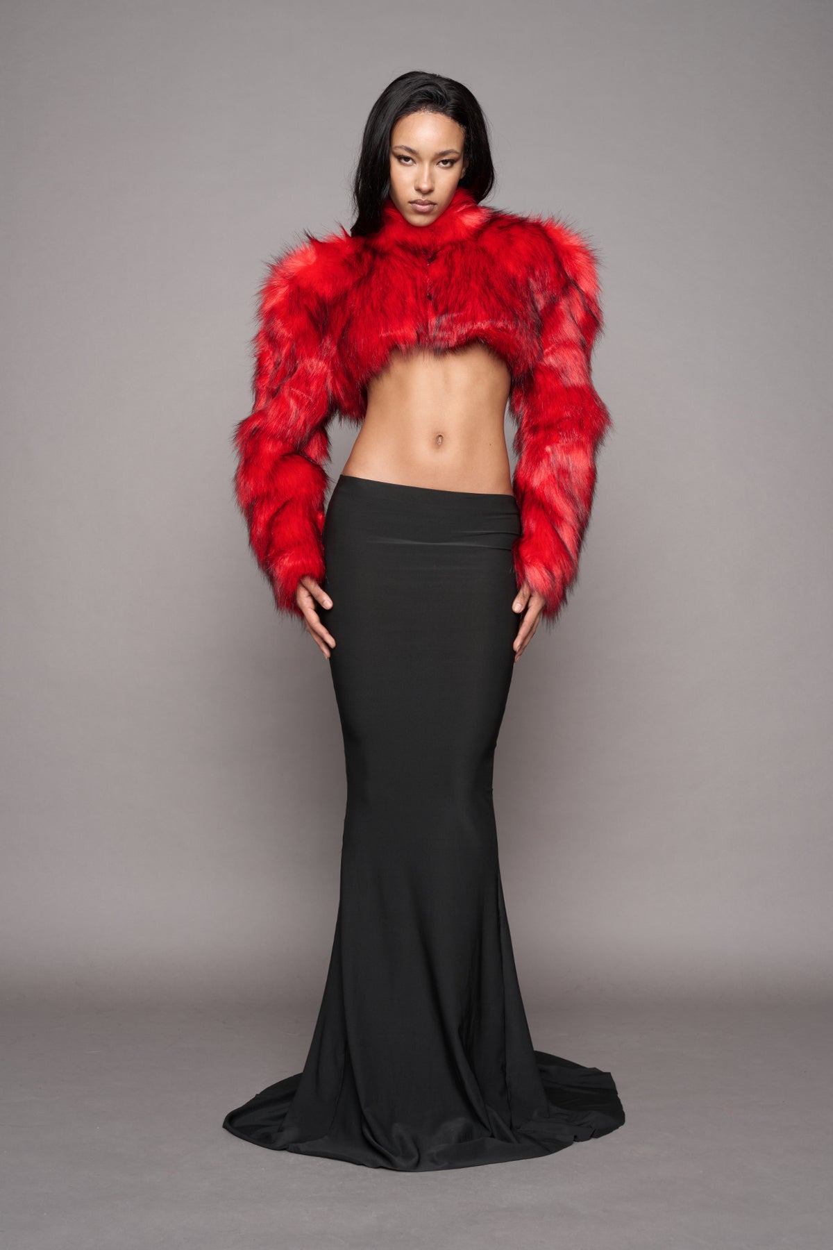 A model stands against a plain gray studio background wearing a bright red faux-fur cropped jacket with long sleeves and a black floor-length mermaid gown.