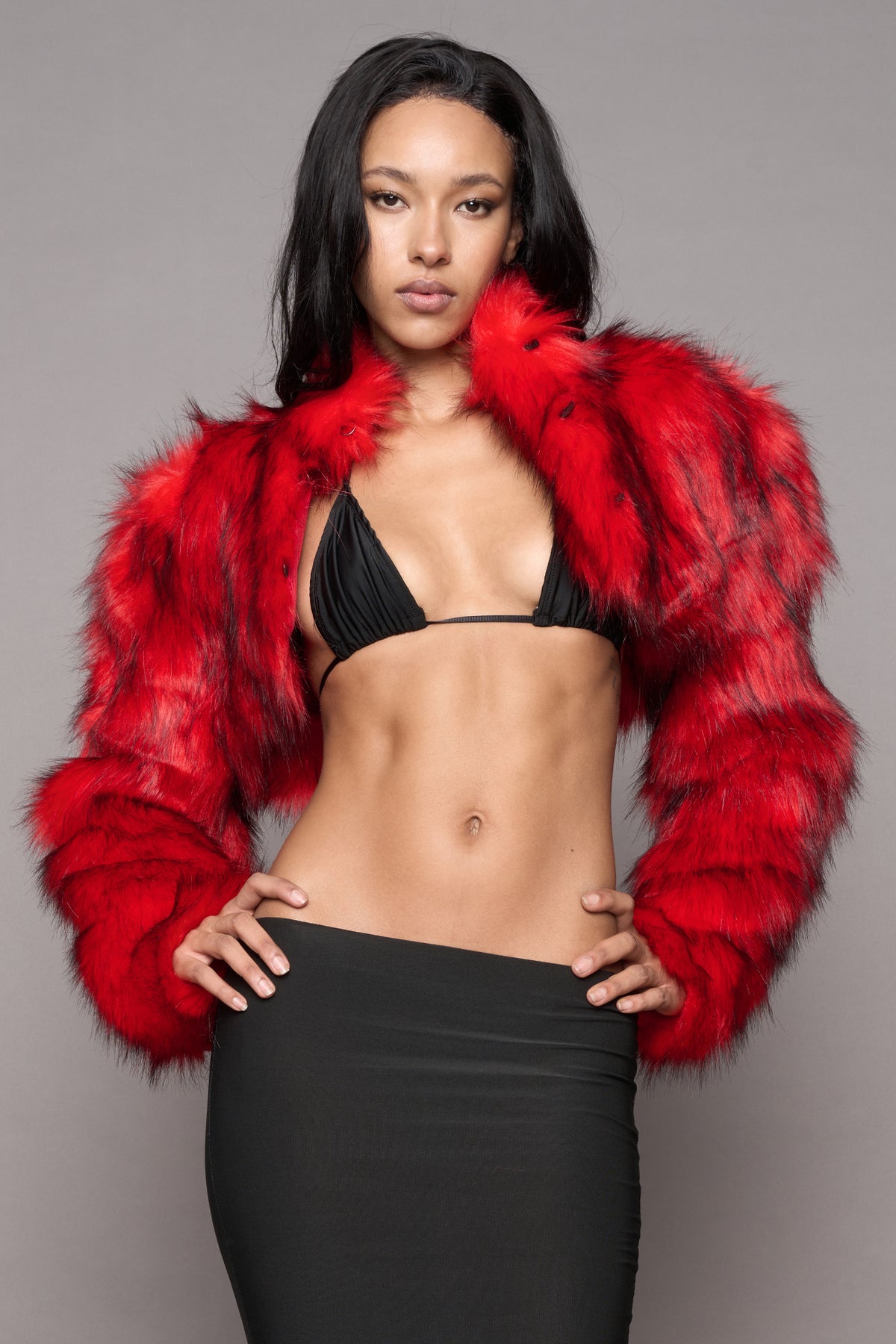 A model posing in a vibrant red faux fur cropped jacket over a black bikini top and a black high-waisted skirt, set against a solid gray studio background.