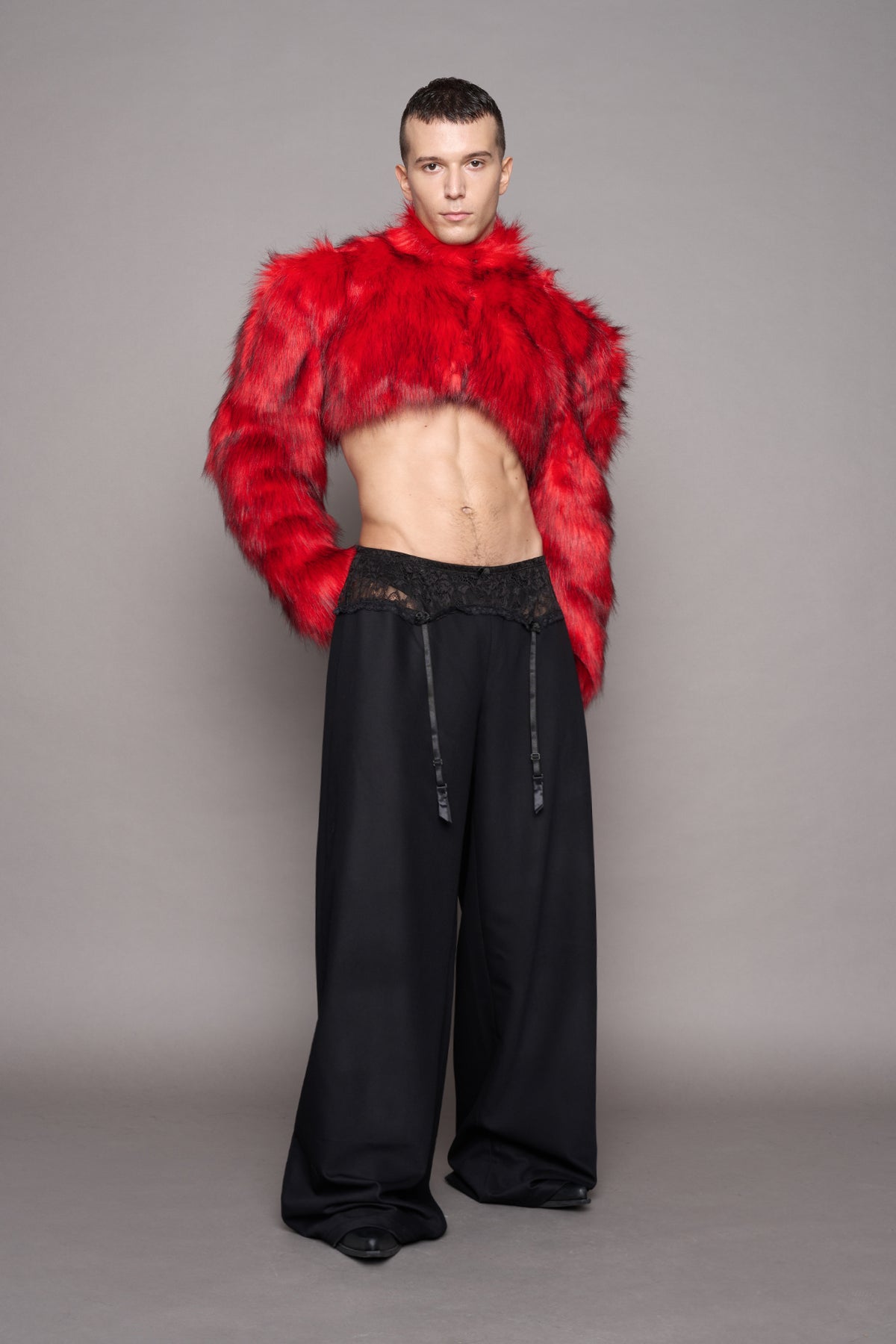 A male model stands against a plain gray studio background wearing a bright red cropped faux-fur jacket over a bare torso, paired with loose black wide-leg pants.
