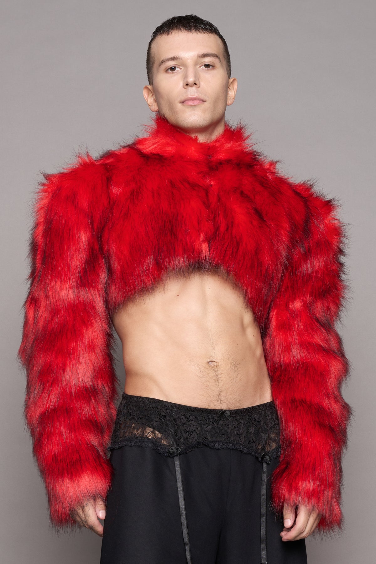 A male model wearing a vibrant red cropped faux fur jacket with long sleeves and a high collar, paired with black pants and a black lace waistband, against a solid gray studio background.
