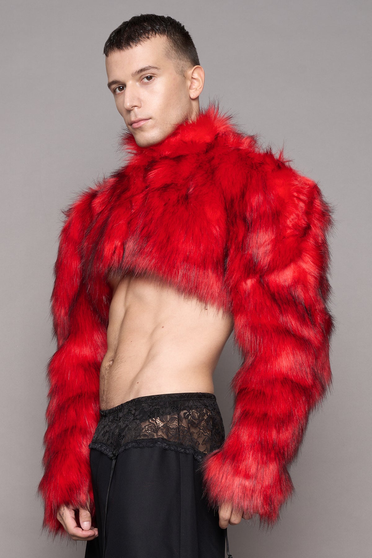 A model wearing a vibrant red cropped faux fur jacket with long sleeves, styled over black pants with a lace waistband, against a solid gray studio background.