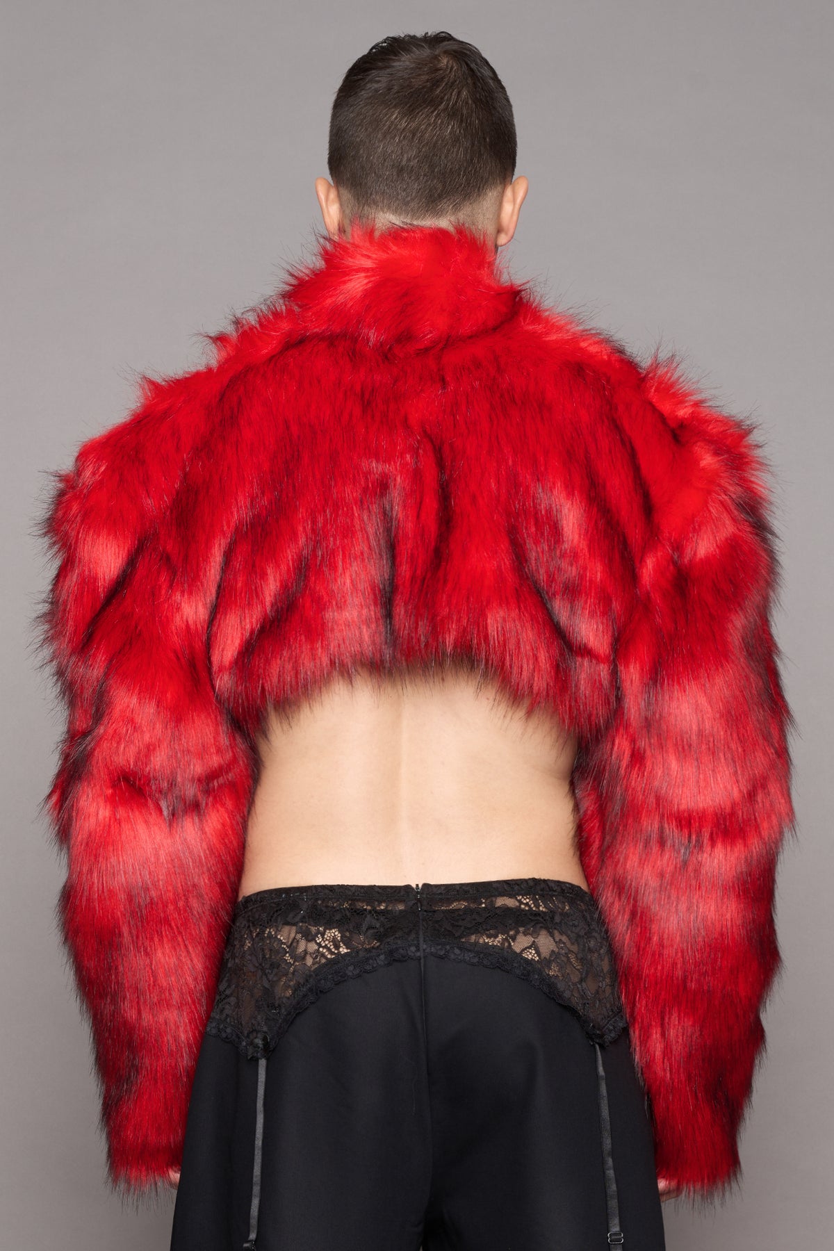 A model shown from the back wearing a vibrant red cropped faux fur jacket and black trousers with a black lace detail against a solid gray studio background.