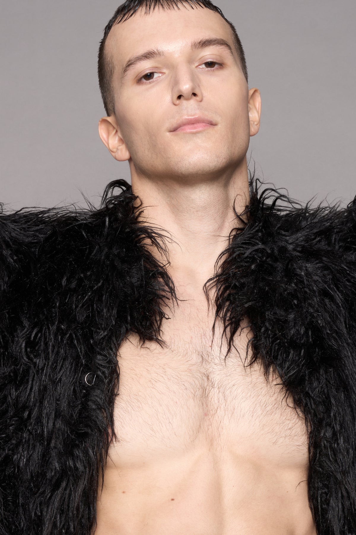A close-up portrait of a man wearing a black shaggy faux-fur jacket, styled open at the chest against a solid gray studio background.