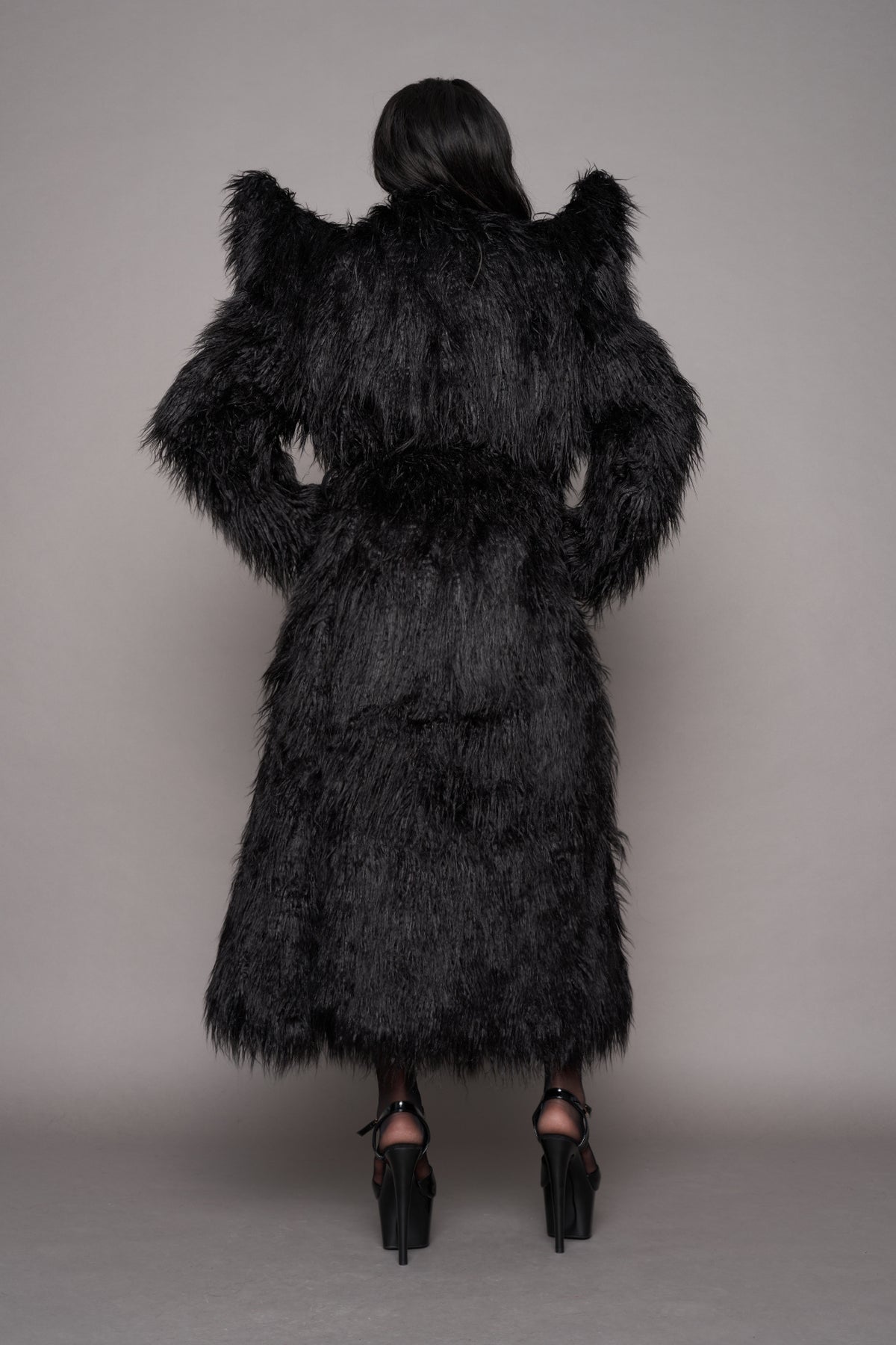 A female model viewed from behind wearing a voluminous black shaggy faux-fur coat with dramatic shoulder spikes, paired with black high heels, against a plain gray studio background.