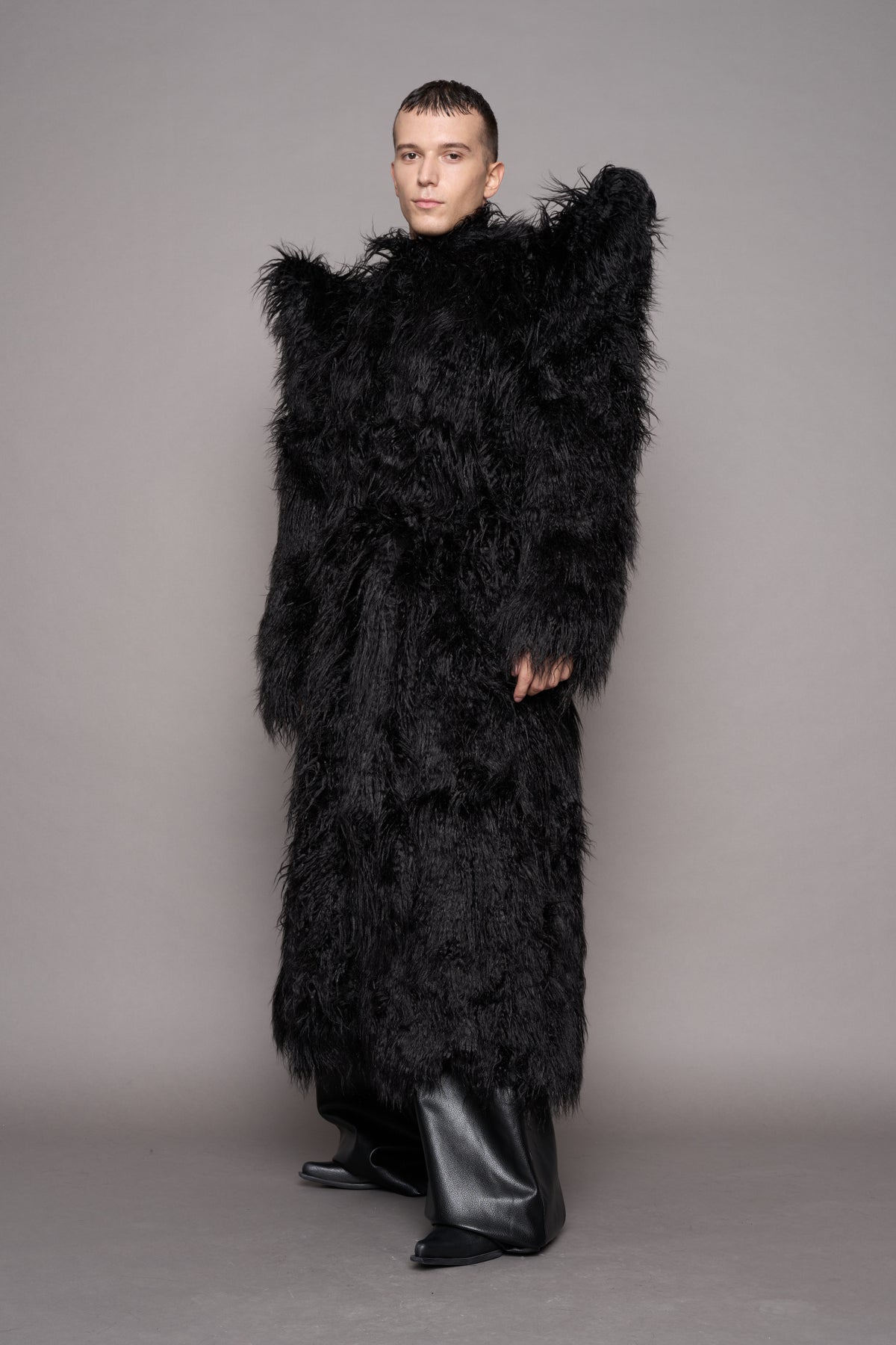 A model wearing a voluminous black shaggy faux-fur coat with dramatic spiky shoulder and black leather boots, standing against a neutral gray studio backdrop.