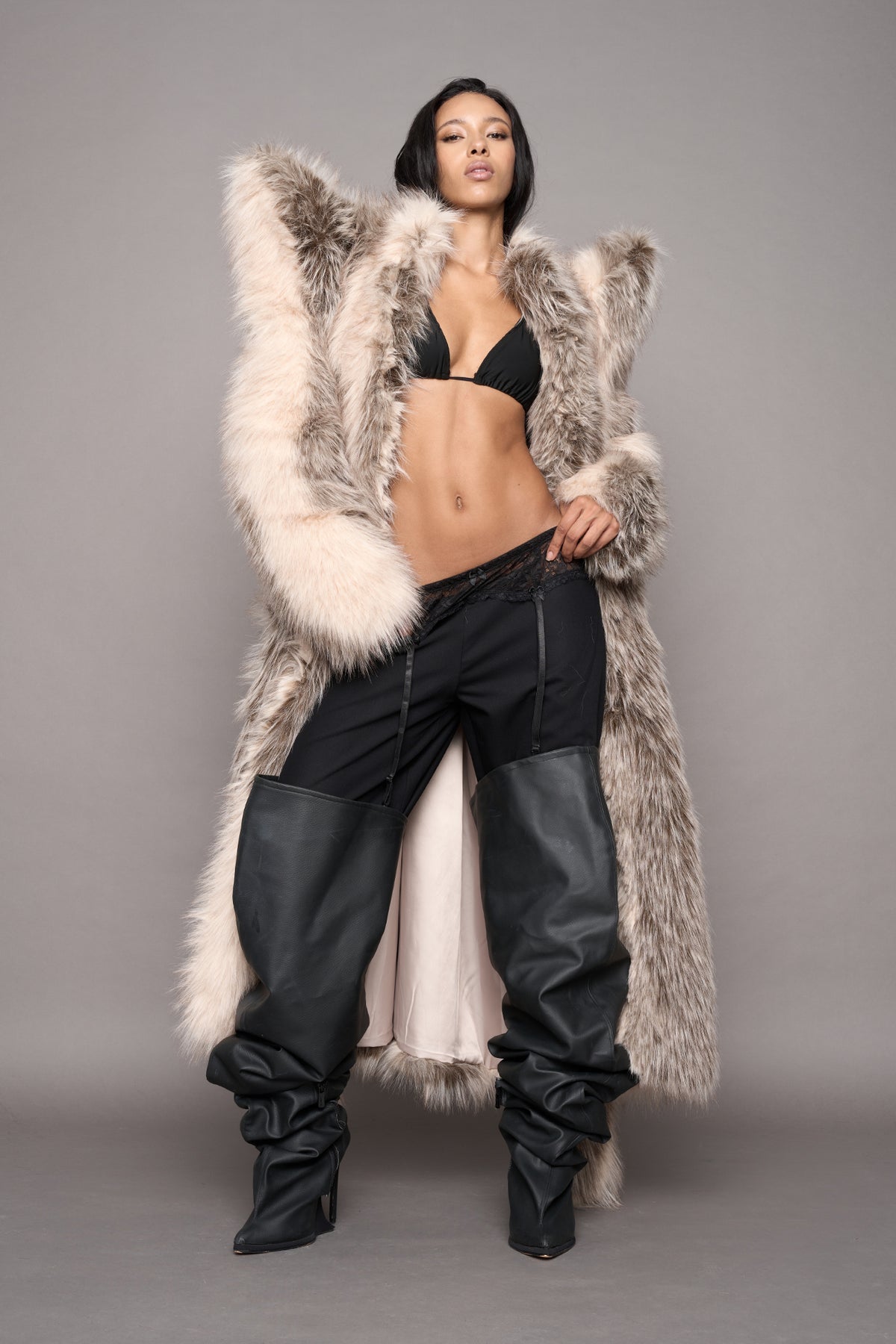 A model poses in a long, oversized black faux-fur coat with spikey shoulders, worn over a black bikini top and black leather pants black leather boots, set against a solid gray studio background.
