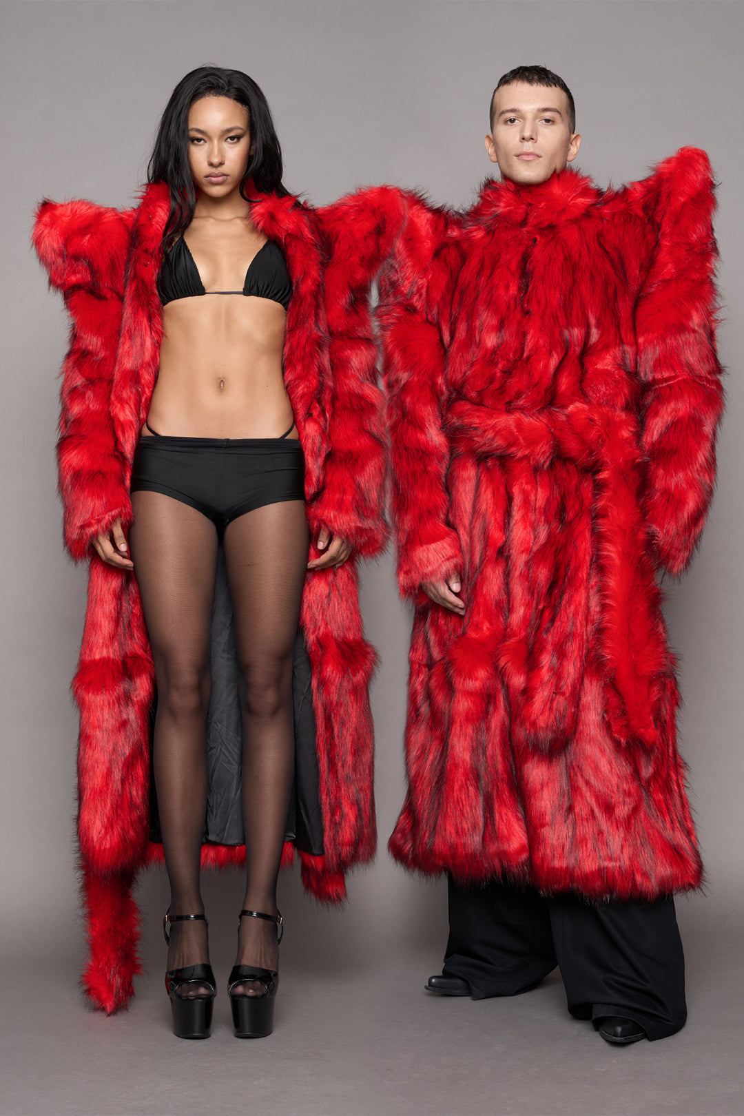 Two models wearing matching oversized faux fur coats in red. The shoulders are wide and spiky. One coat is worn open styled with a bikini top and short and heels, he over coat is closed with a matching fur red waist tie and black pants