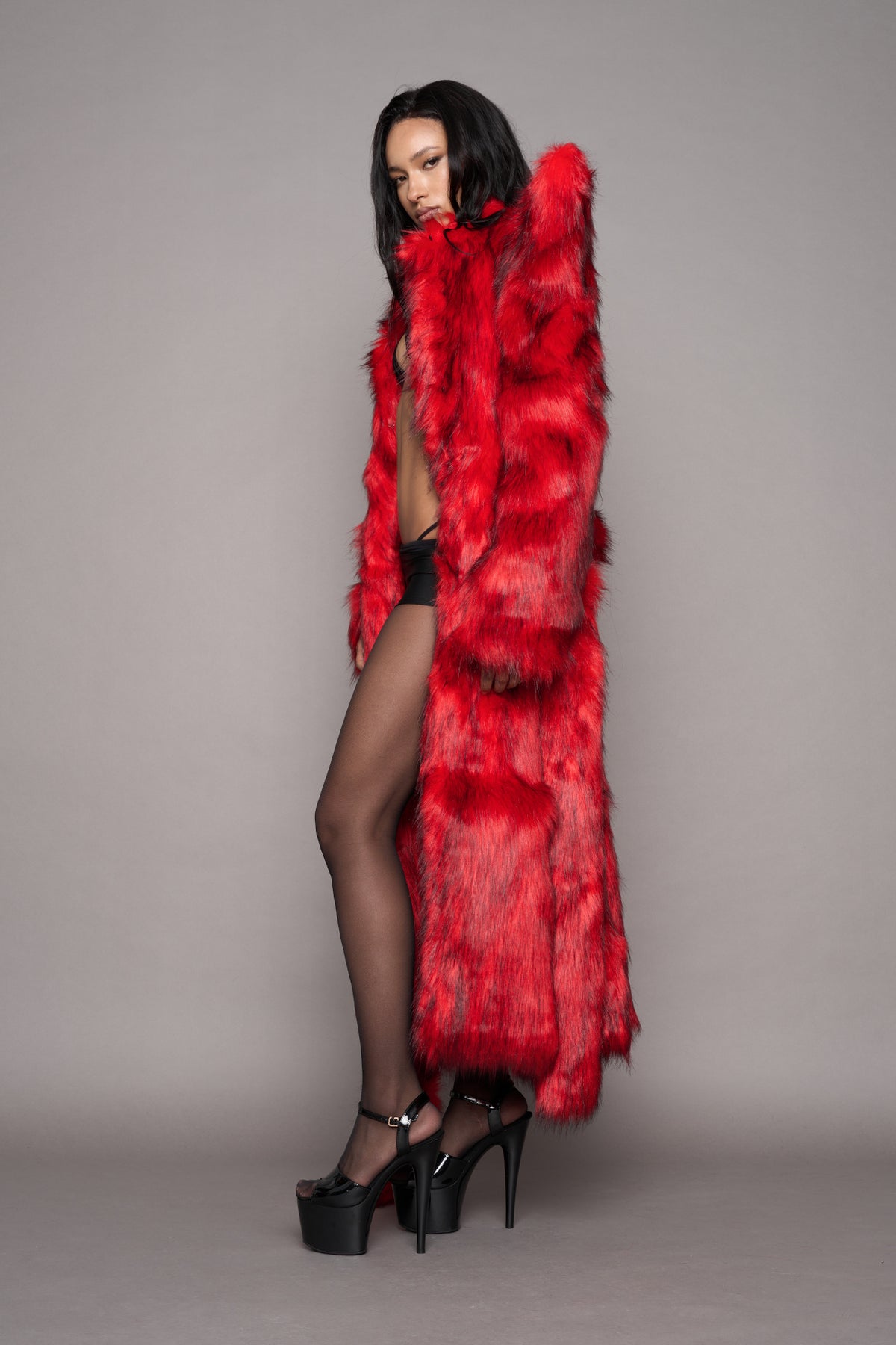 A model wearing a vibrant red faux fur coat with big spiky shoulders, sheer black tights, and black high-heeled platform shoes, posed against a solid gray studio background.
