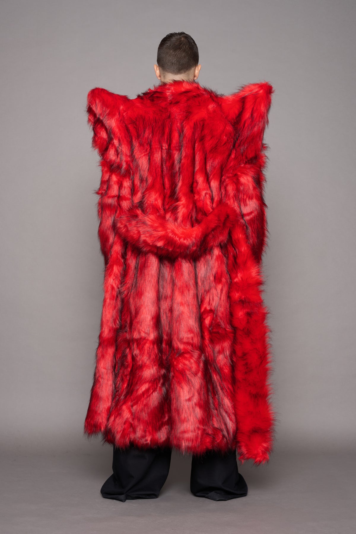 A model shown from the back wearing a voluminous, bright red shaggy faux-fur coat with dramatic shoulder spikes, paired with black trousers, against a solid gray studio background.