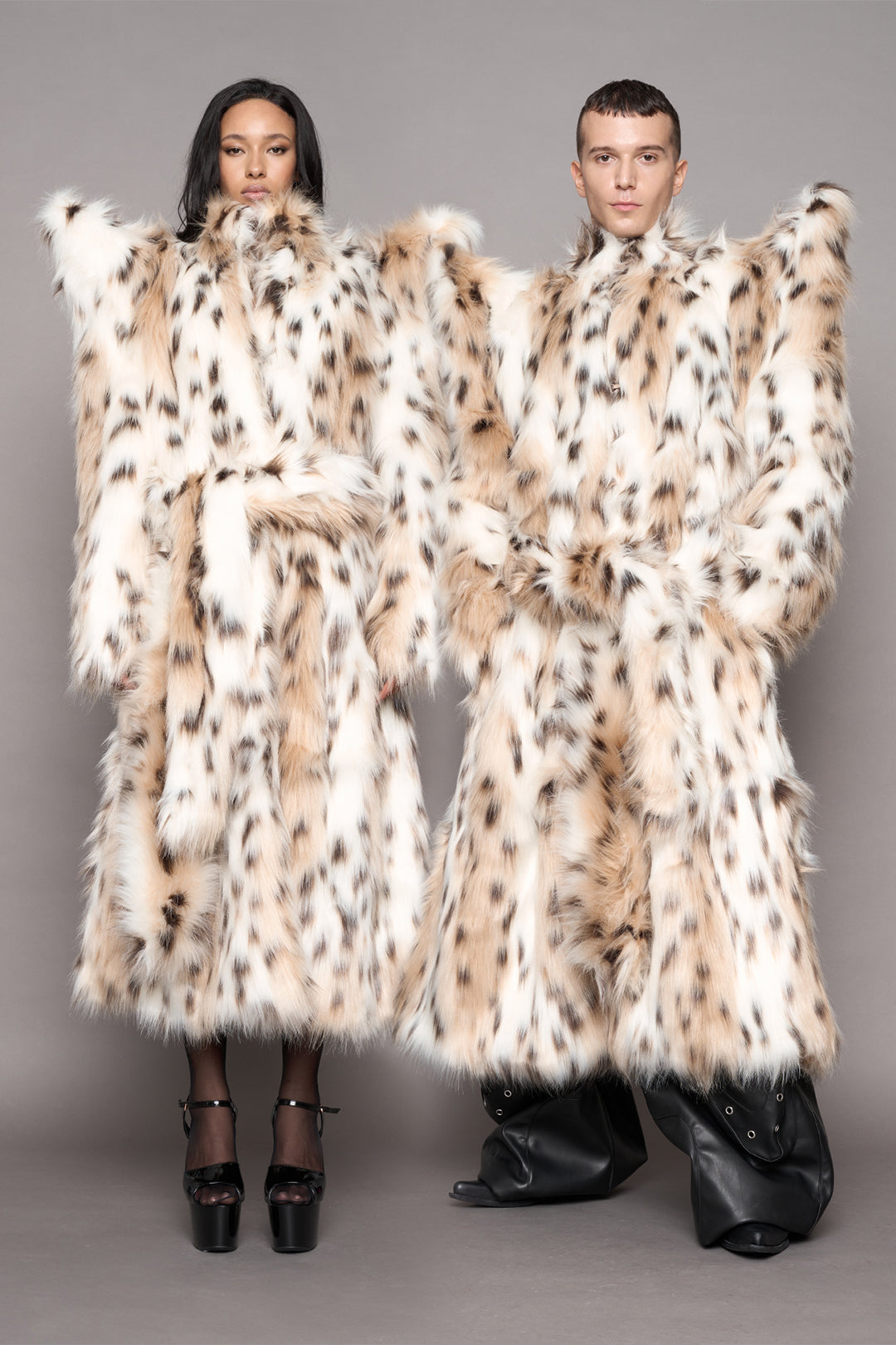 Two models wearing oversized, high-volume faux fur coats with a leopard pattern, posed against a solid gray studio background.