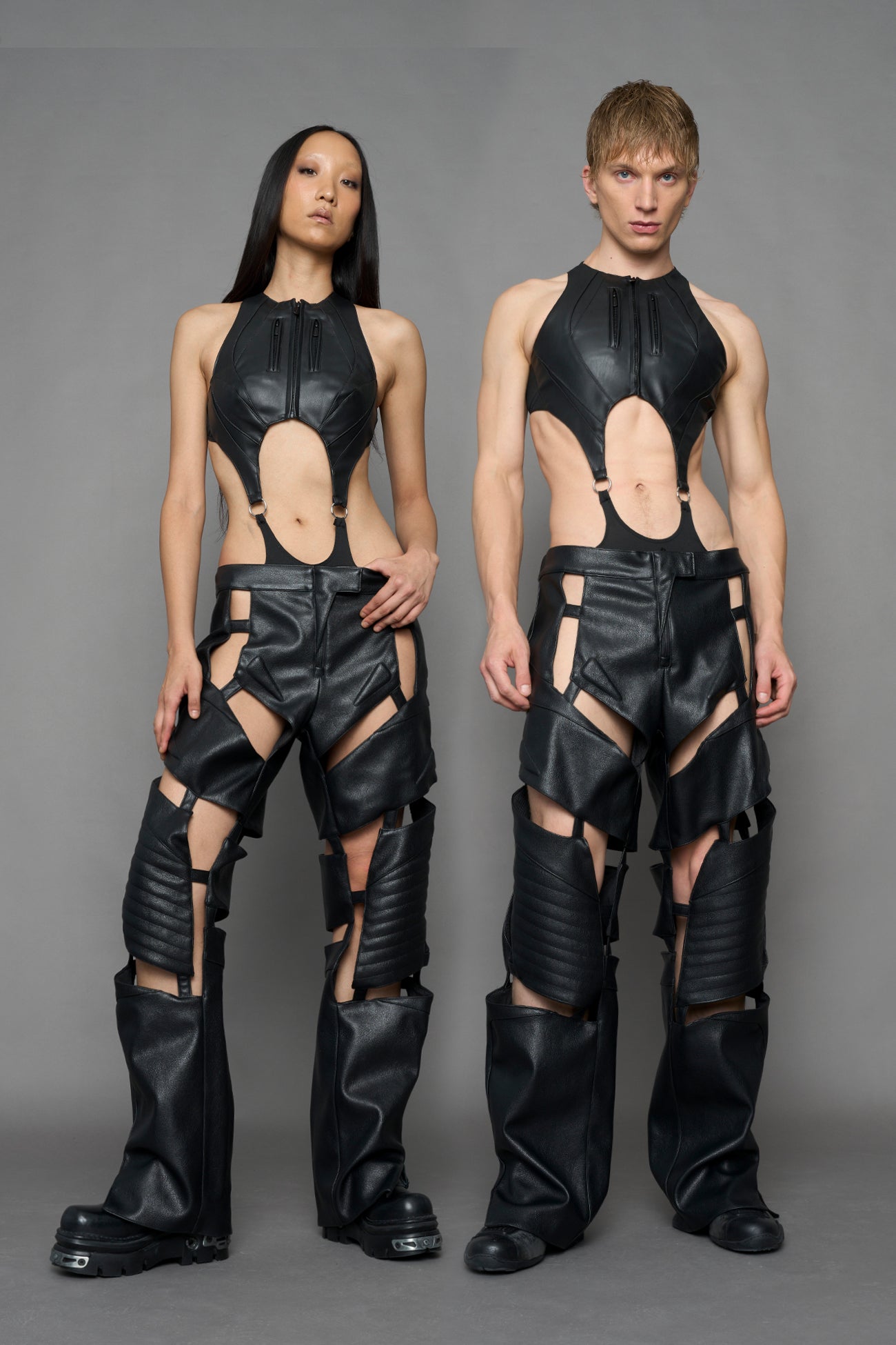 A man and a woman modeling matching futuristic black faux-leather outfits with dramatic cutouts, standing against a plain gray studio background.