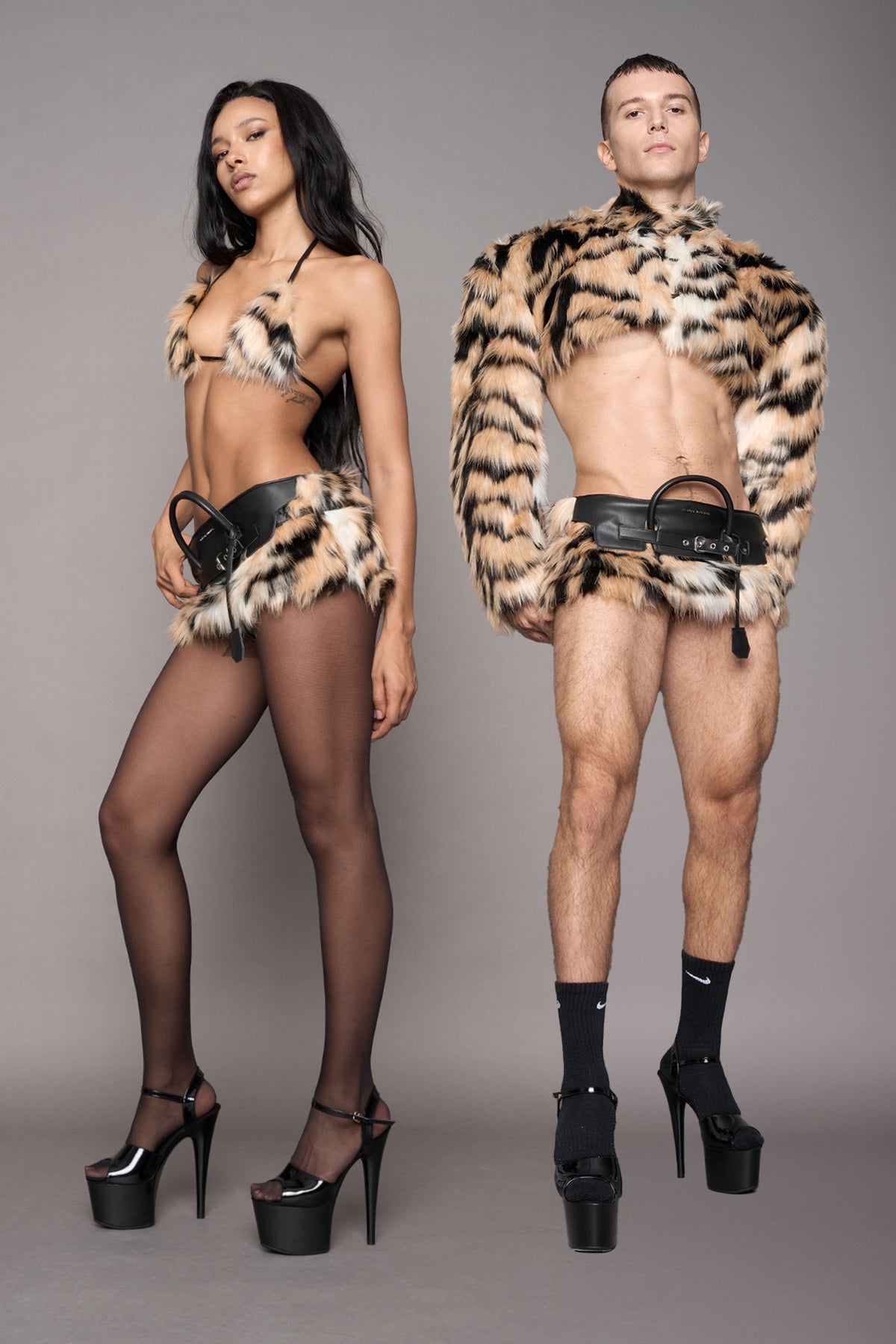 Two models posing in tiger faux fur miniskirt against a solid gray studio background. The female model wears a leopard-print halter bikini top and matching mini skirt with black thigh-high stockings and platform heels and the male model is wearing a tiger fur matching cropped bolero jacket