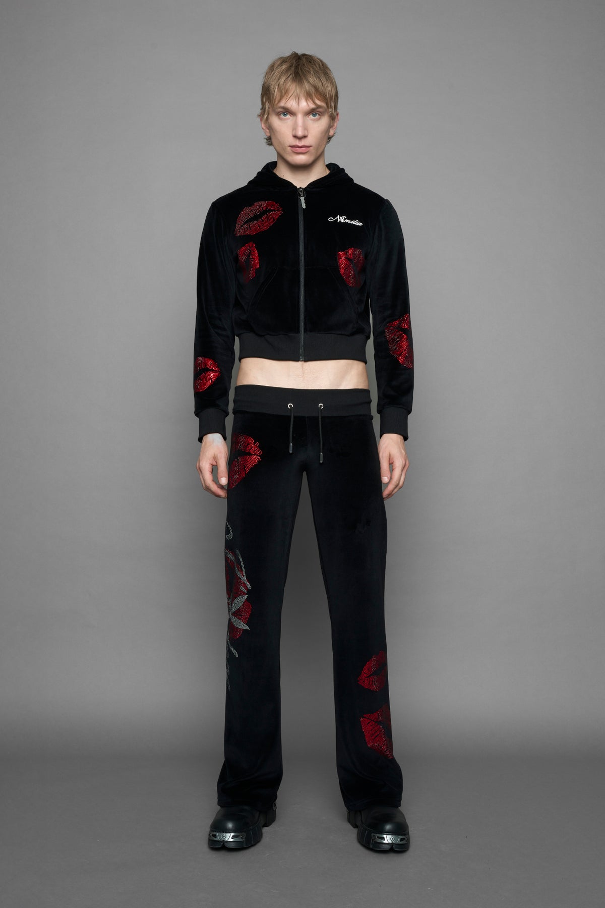A female model wearing a black velvet zip-up hoodie with red sequined lip-print patches on the chest and sleeve, featuring white 'Namilia' embroidery on the left chest, photographed against a solid gray studio background.
