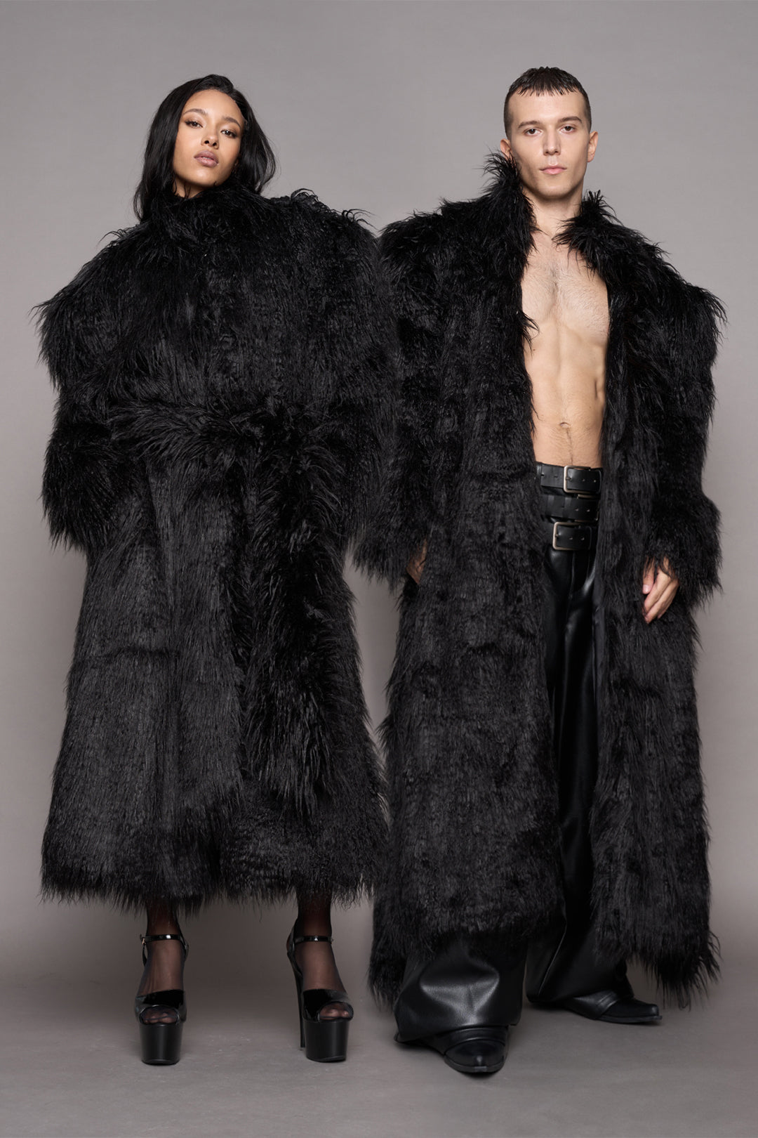 A man and a woman modeling matching oversized black shaggy faux-fur coats against a plain gray studio background.