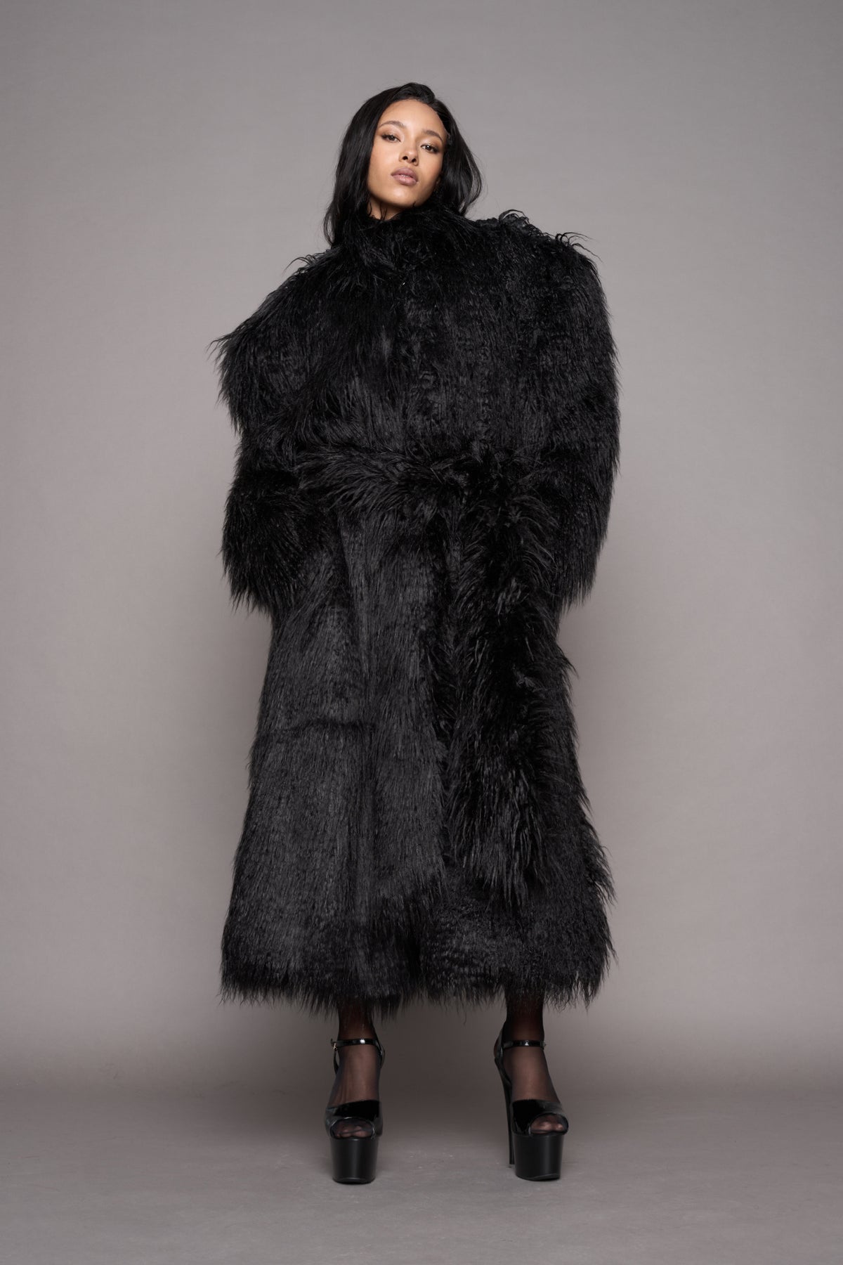 A female model wearing a voluminous, oversized black shaggy faux-fur coat with a high collar and a matching waist tie, styled with black platform heels, against a solid gray studio background.