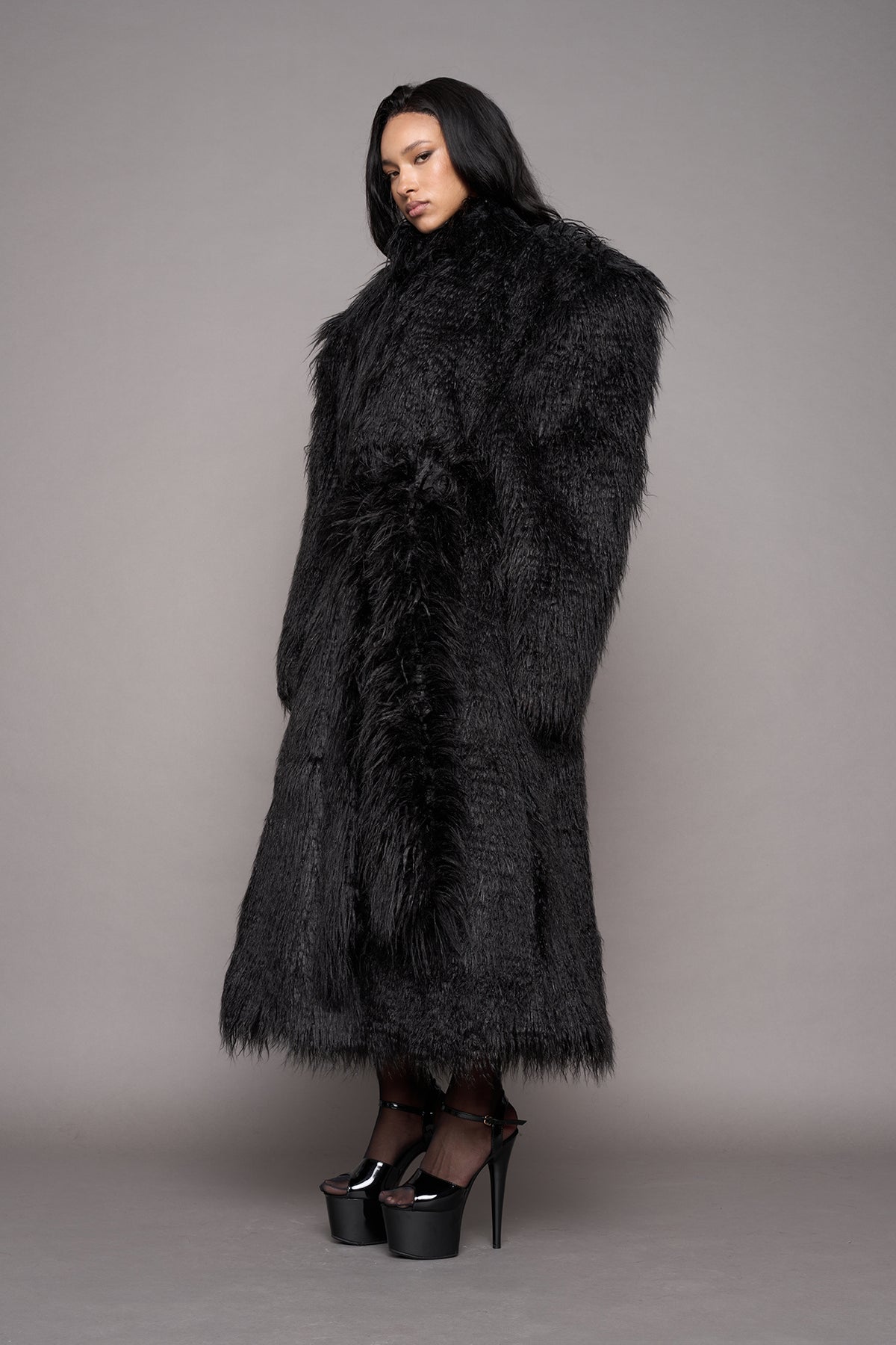 A model wearing a voluminous, oversized black faux fur coat with a high collar and a matching waist tie, styled with black platform heels against a solid gray studio background.