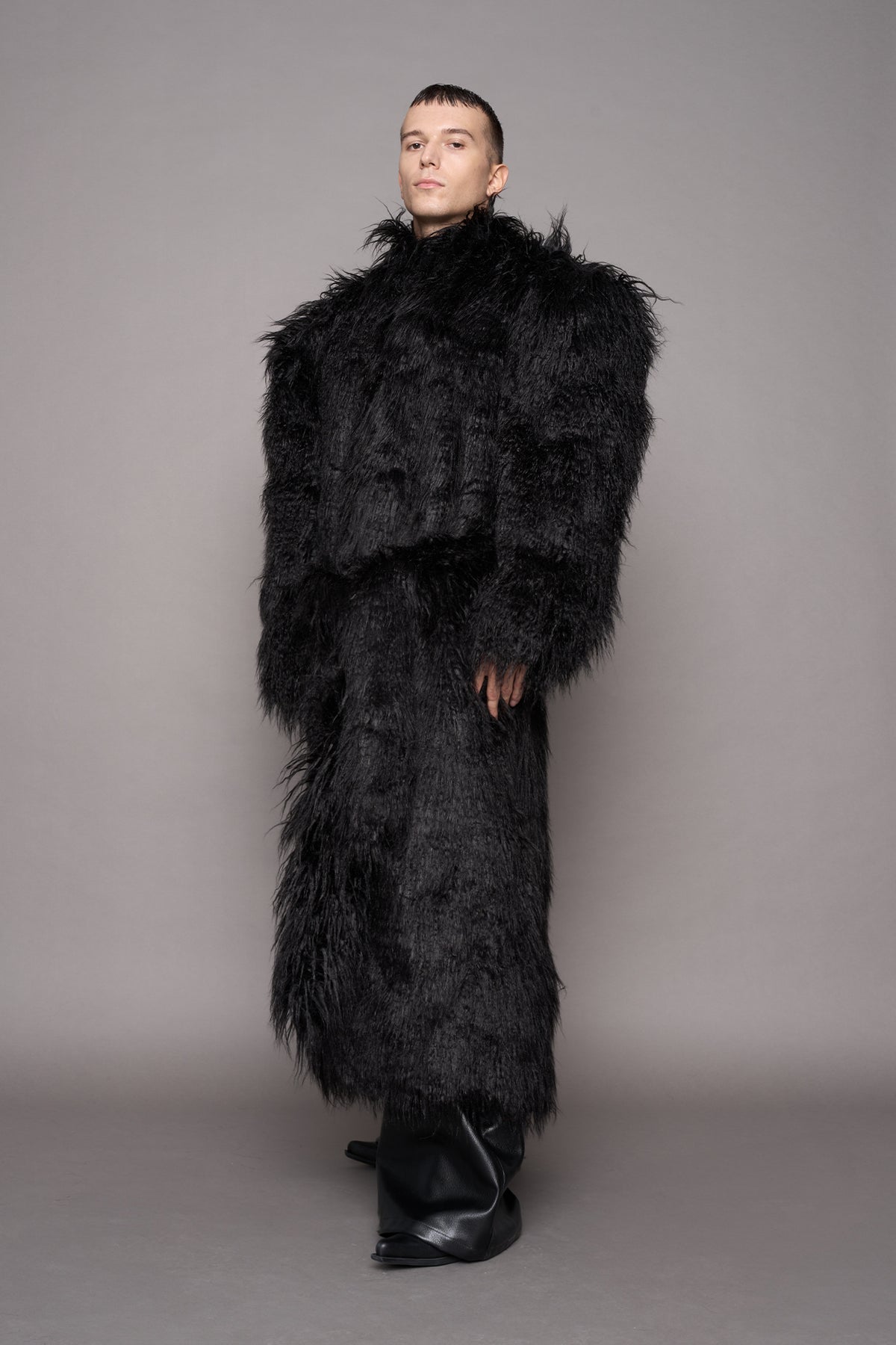A model wearing a voluminous black shaggy faux-fur coat and matching waist tie, paired with black pointed boots, photographed against a neutral gray studio backdrop.