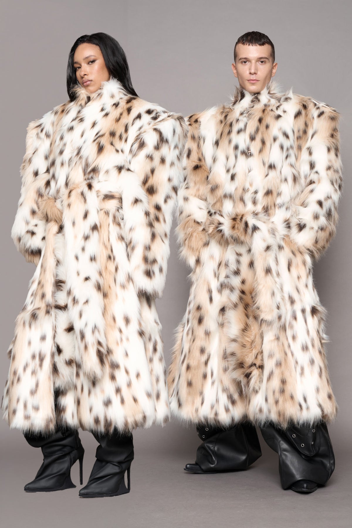 Two models wearing matching oversized faux-fur coats with leopard pattern, paired with black boots, against a solid gray studio background.