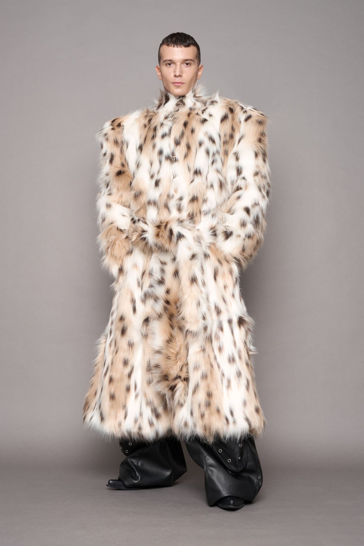 A model wearing a voluminous, oversized faux-fur coat with a beige, brown, and black spotted leopard pattern, paired with black boots, against a solid gray studio background.