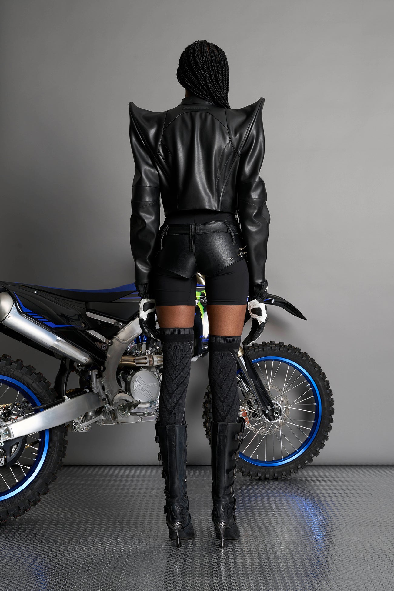 Model wears the black Namilia Spike Moto Jacket with wire shoulder pads, branded zipper, band collar, zippered breast and waist pockets, motocross-style lines, and black logo rubber tag on the back.
