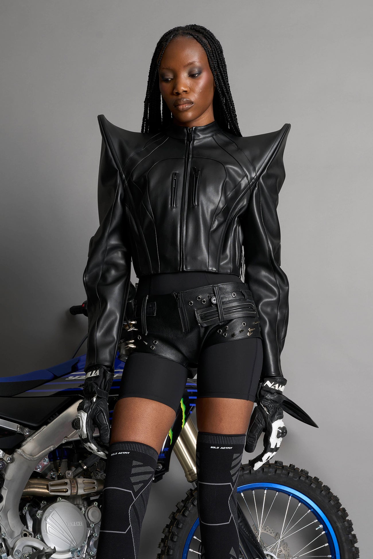 Model wears the black Namilia Spike Moto Jacket with wire shoulder pads, branded zipper, band collar, zippered breast and waist pockets, motocross-style lines, and black logo rubber tag on the back.