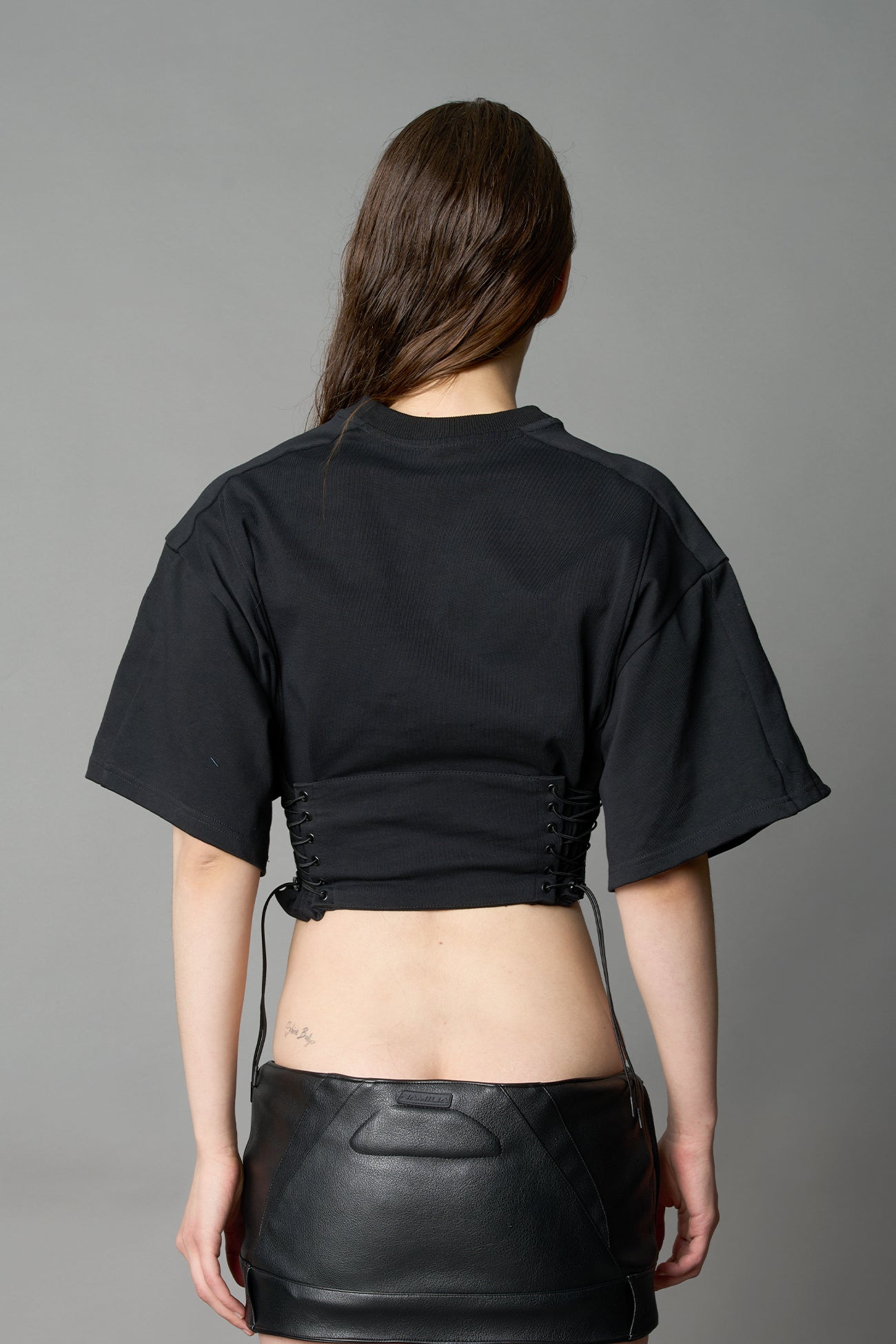 Front view of model in black Namilia Corset-T shirt with sculpted corset-style waist, curved hemline, and oversized sleeves, styled with leather mini skirt and moto boots.