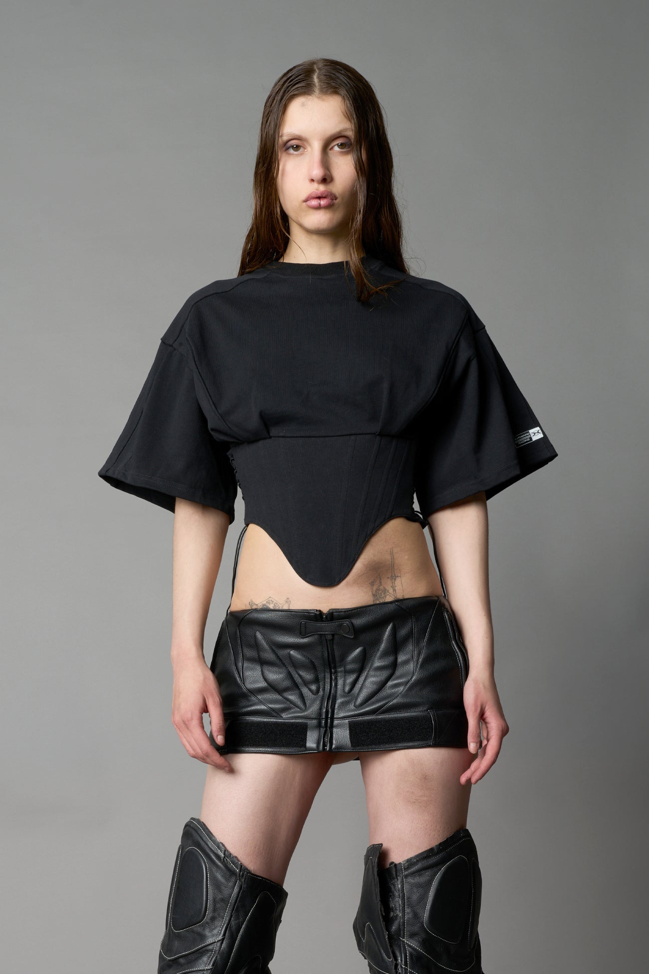 Front view of model in black Namilia Corset-T shirt with sculpted corset-style waist, curved hemline, and oversized sleeves, styled with leather mini skirt and moto boots.