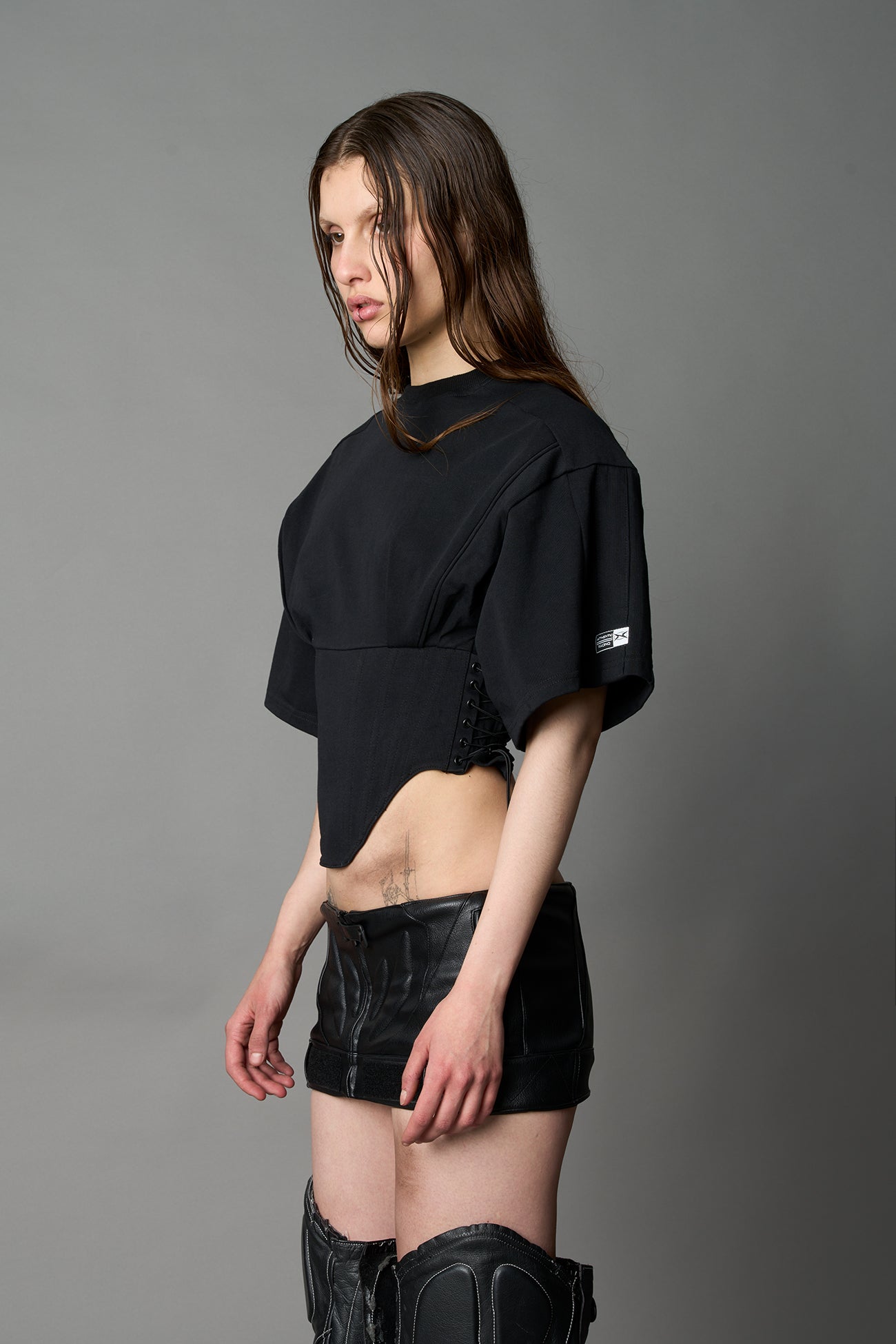 Front view of model in black Namilia Corset-T shirt with sculpted corset-style waist, curved hemline, and oversized sleeves, styled with leather mini skirt and moto boots.