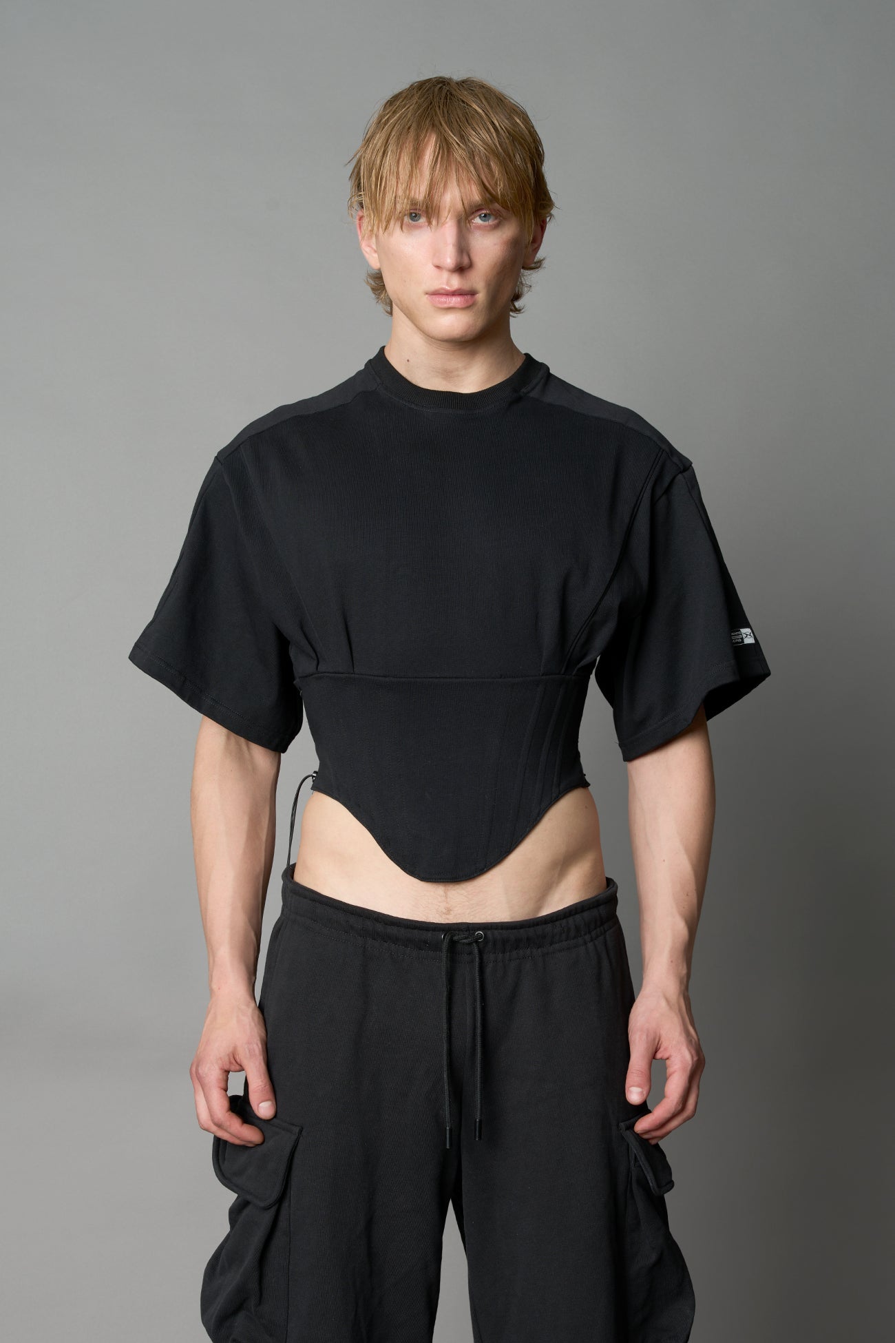 Front view of model in black Namilia Corset-T shirt with sculpted corset-style waist, curved hemline, and oversized sleeves, styled with leather mini skirt and moto boots.
