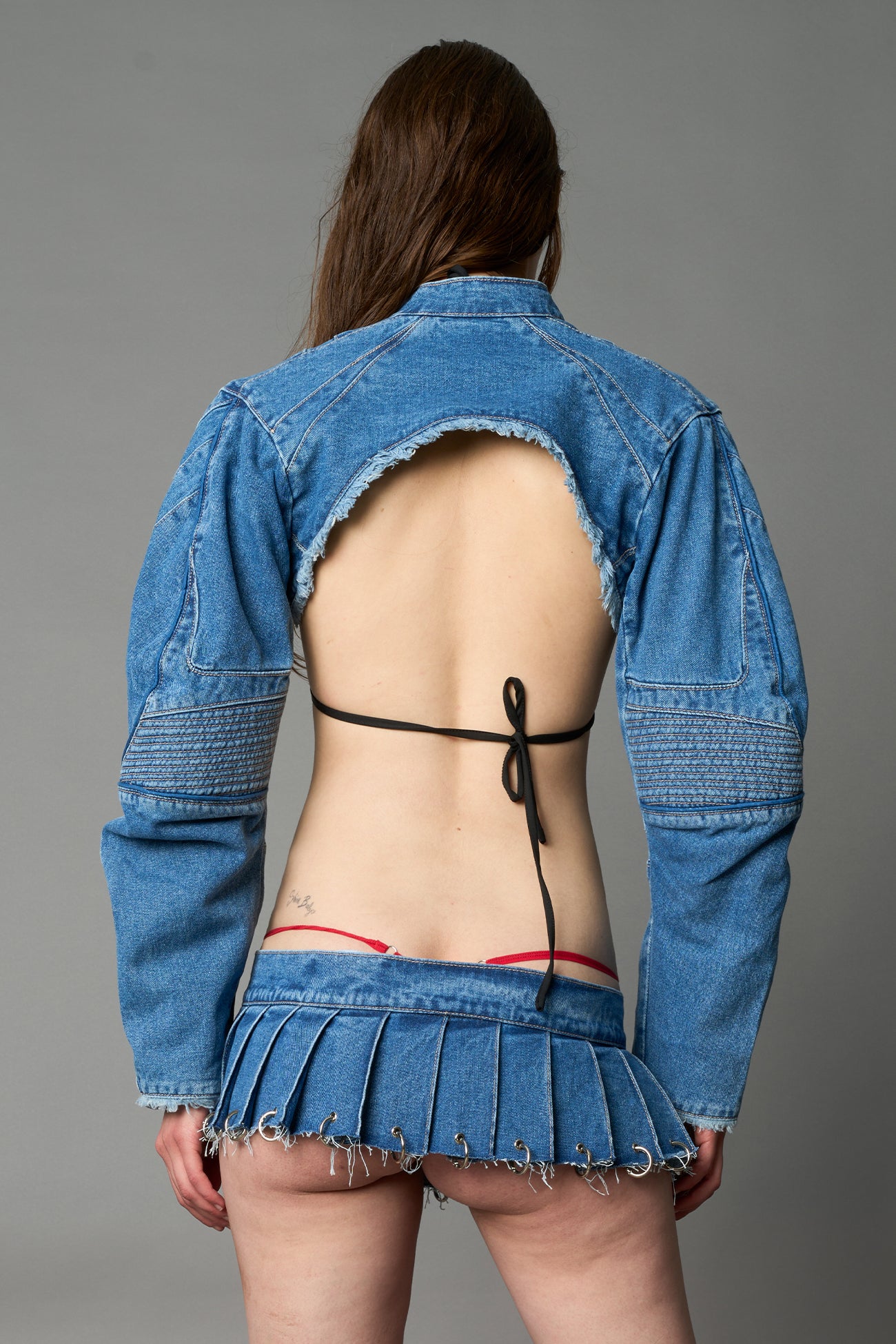 model in ultra-short blue washed denim pleated mini skirt featuring frayed hem, silver grommet rings, and wide belt-style front closure
