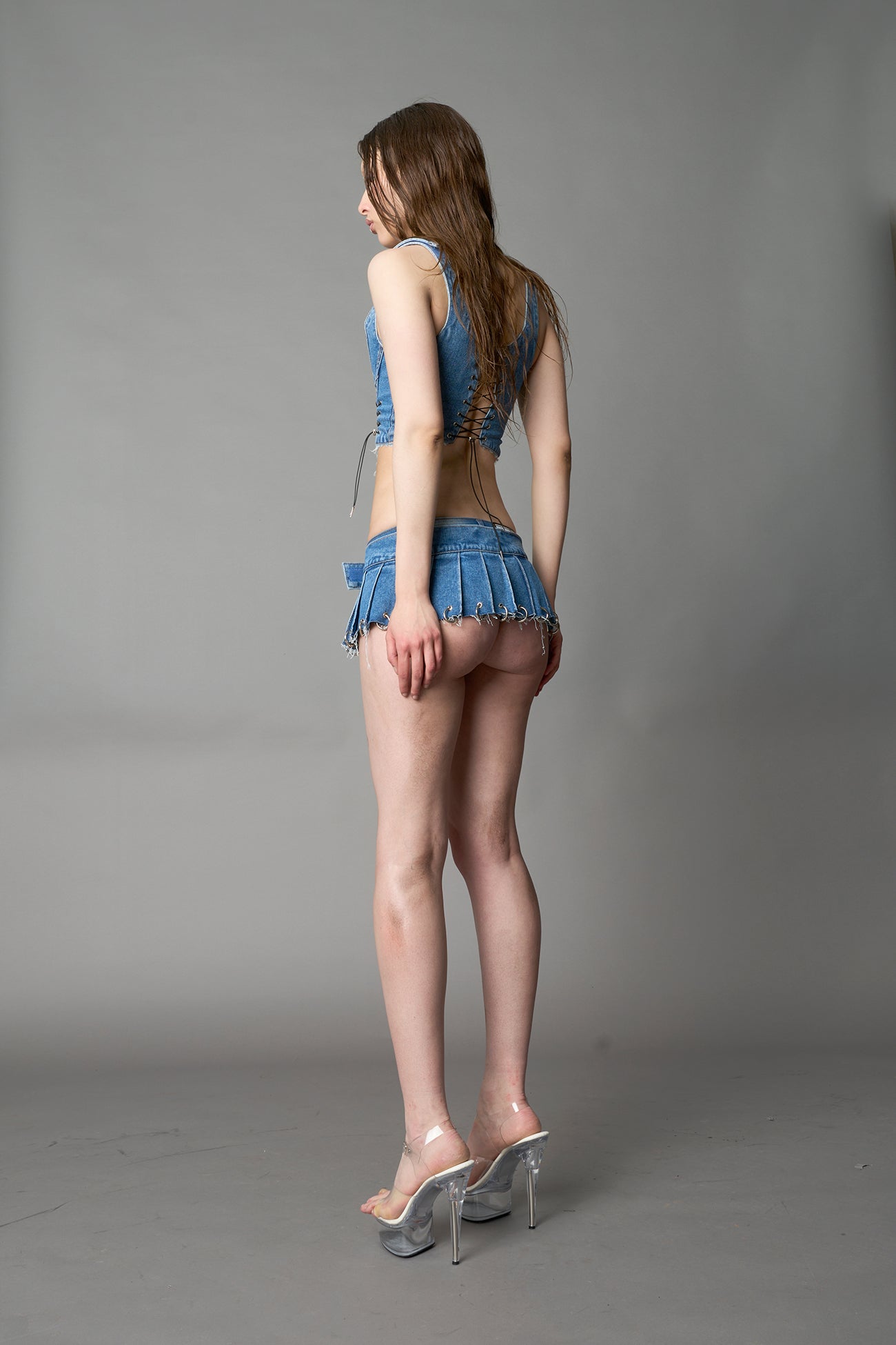 model in ultra-short blue washed denim pleated mini skirt featuring frayed hem, silver grommet rings, and wide belt-style front closure