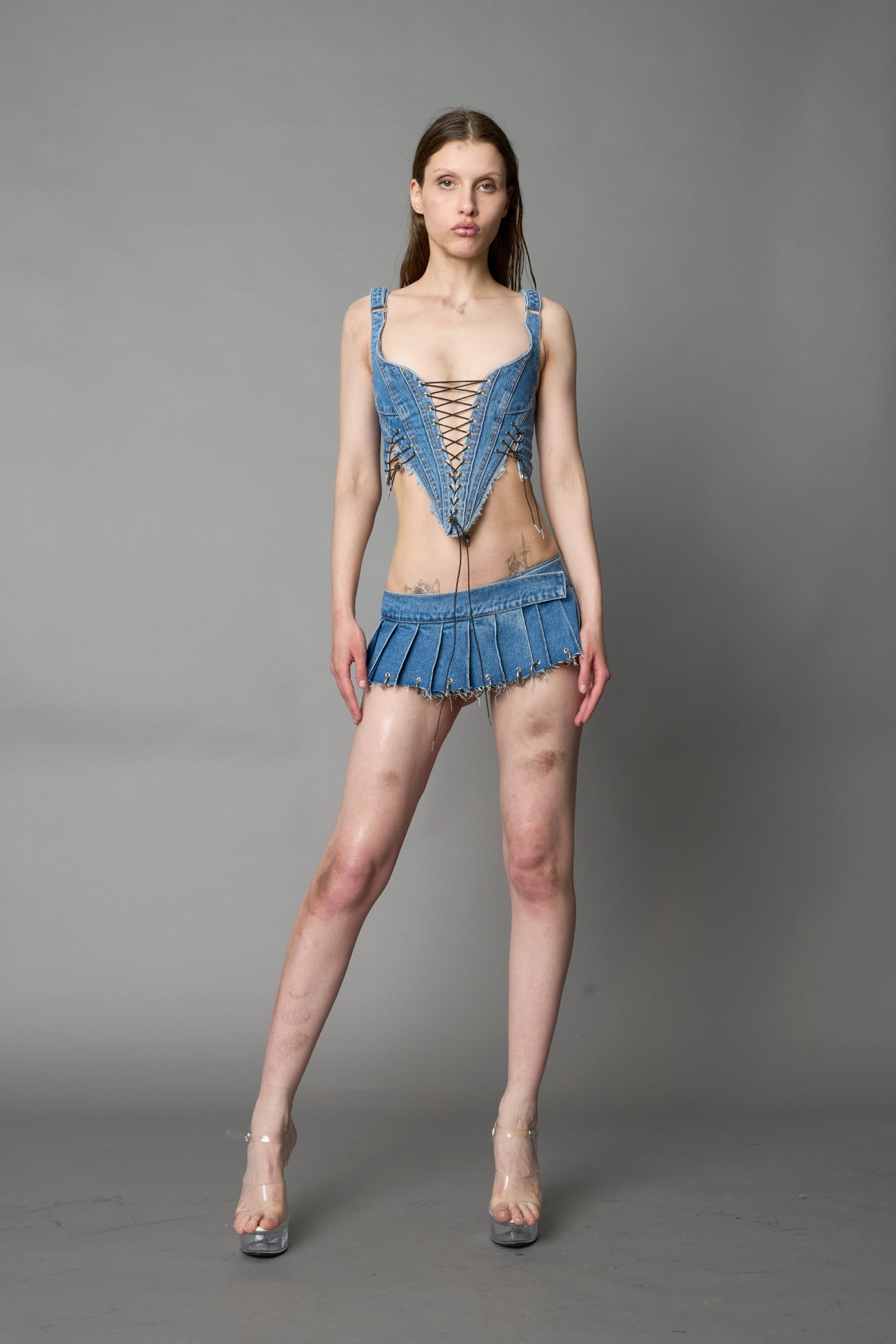 model in ultra-short blue washed denim pleated mini skirt featuring frayed hem, silver grommet rings, and wide belt-style front closure