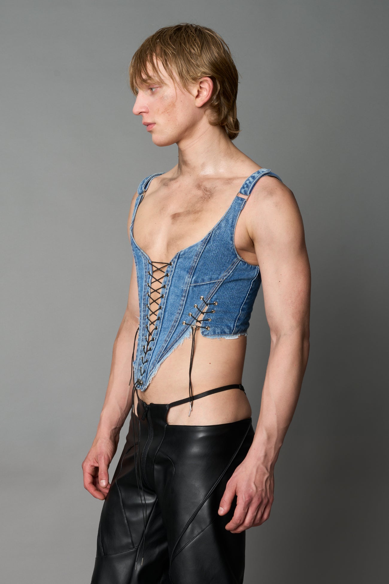 Model wearing blue washed denim corset top with plunging neckline, lace-up front and sides, raw frayed hem, and structured fit