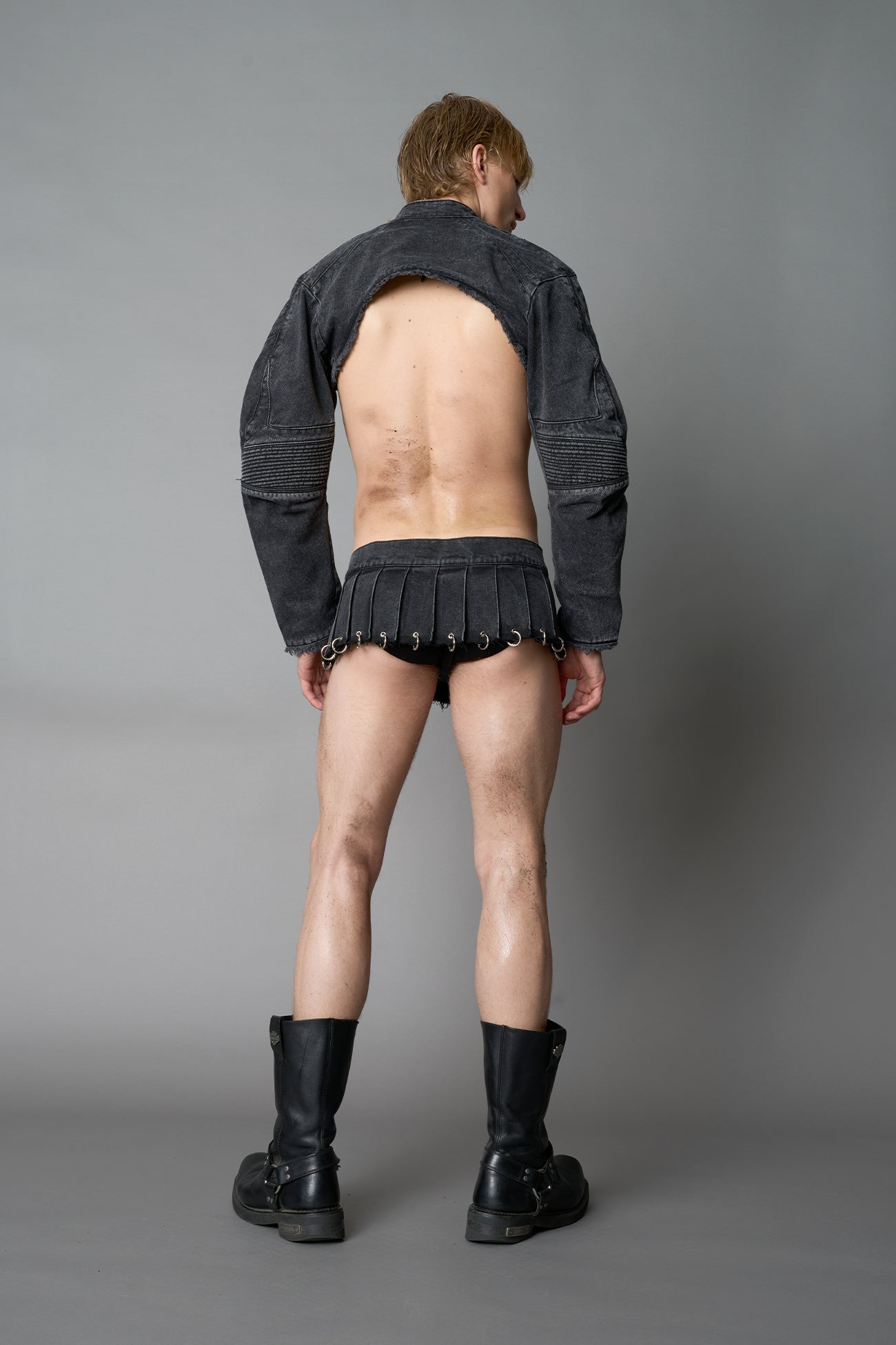 model in ultra-short black washed denim pleated mini skirt featuring frayed hem, silver grommet rings, and wide belt-style front closure