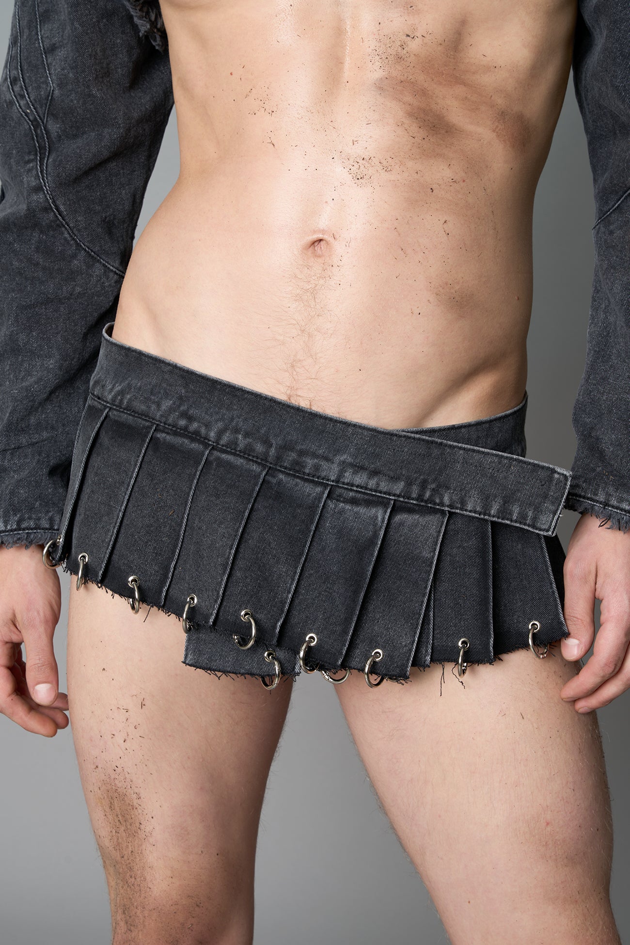 model in ultra-short black washed denim pleated mini skirt featuring frayed hem, silver grommet rings, and wide belt-style front closure