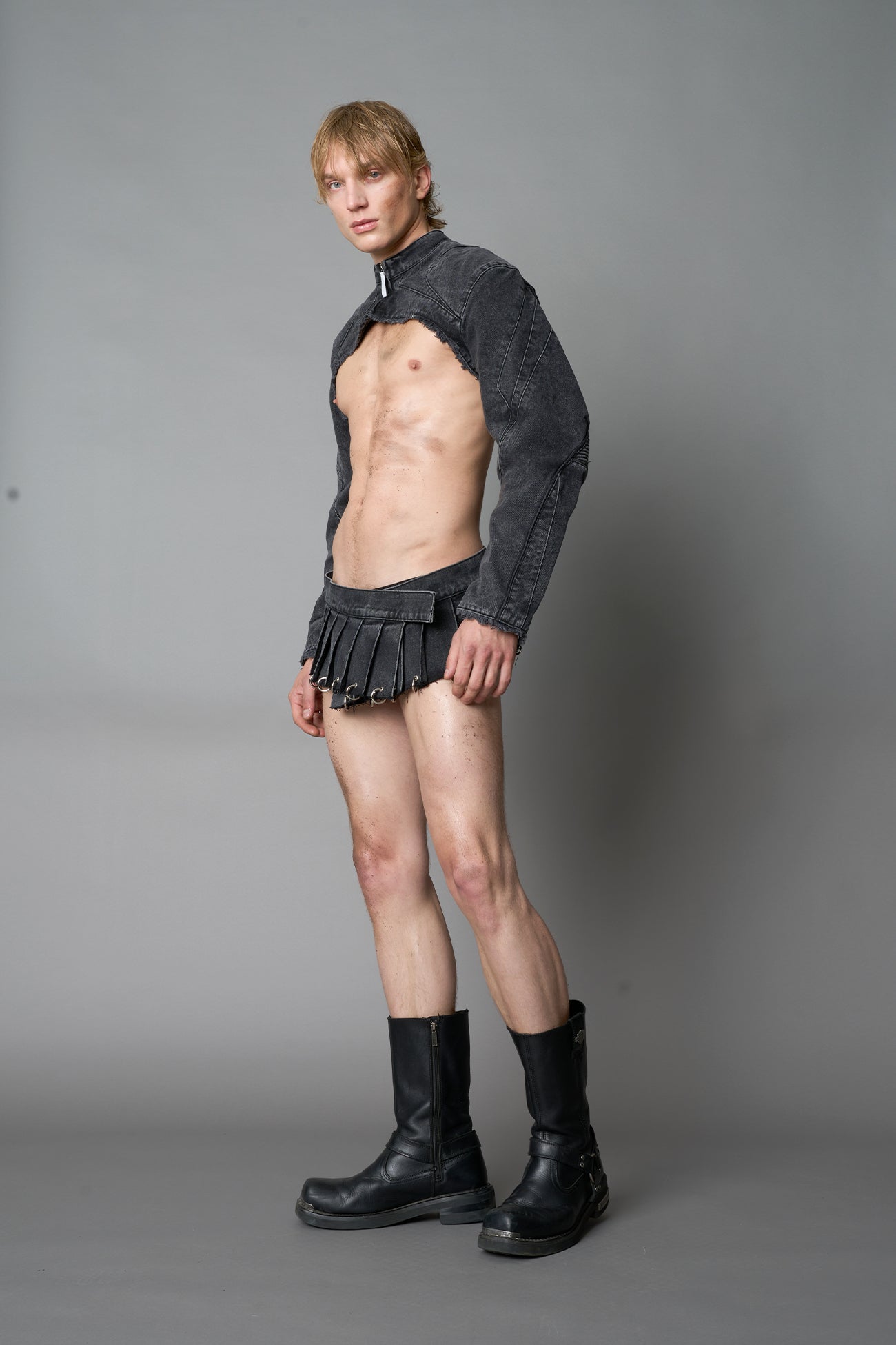 model in ultra-short black washed denim pleated mini skirt featuring frayed hem, silver grommet rings, and wide belt-style front closure
