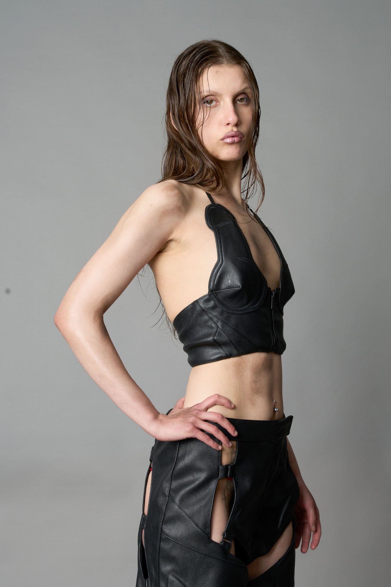 Model in bold black vegan leather statement halter top with provocative cup design and front zipper, paired with matching pants – futuristic streetwear look
