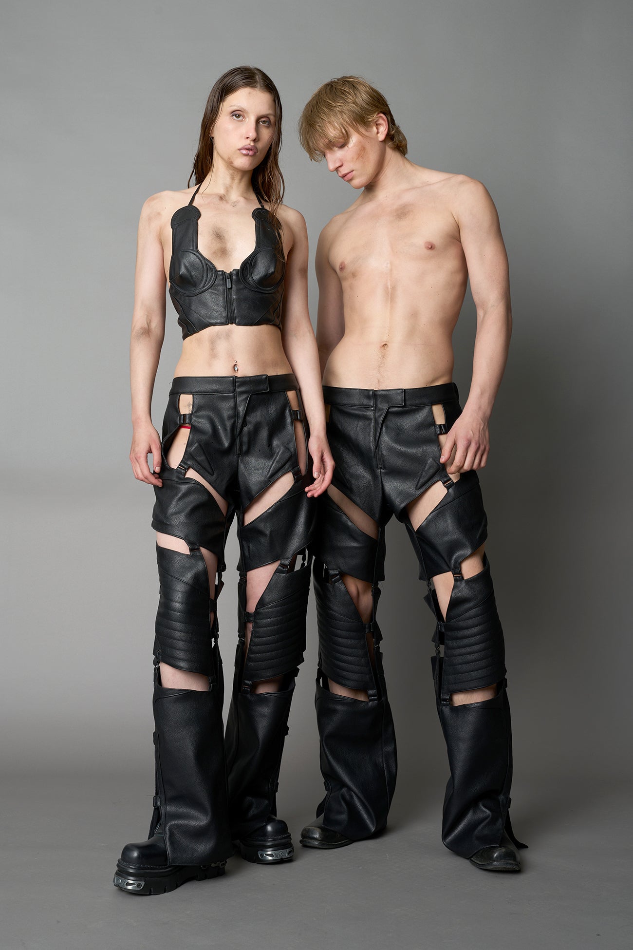 Model wearing black vegan leather motocross-inspired pants with thigh cut-outs, strap harness details, quilted knee padding, and full-length leg gaiters; styled with a structured black bandeau top against a neutral background.