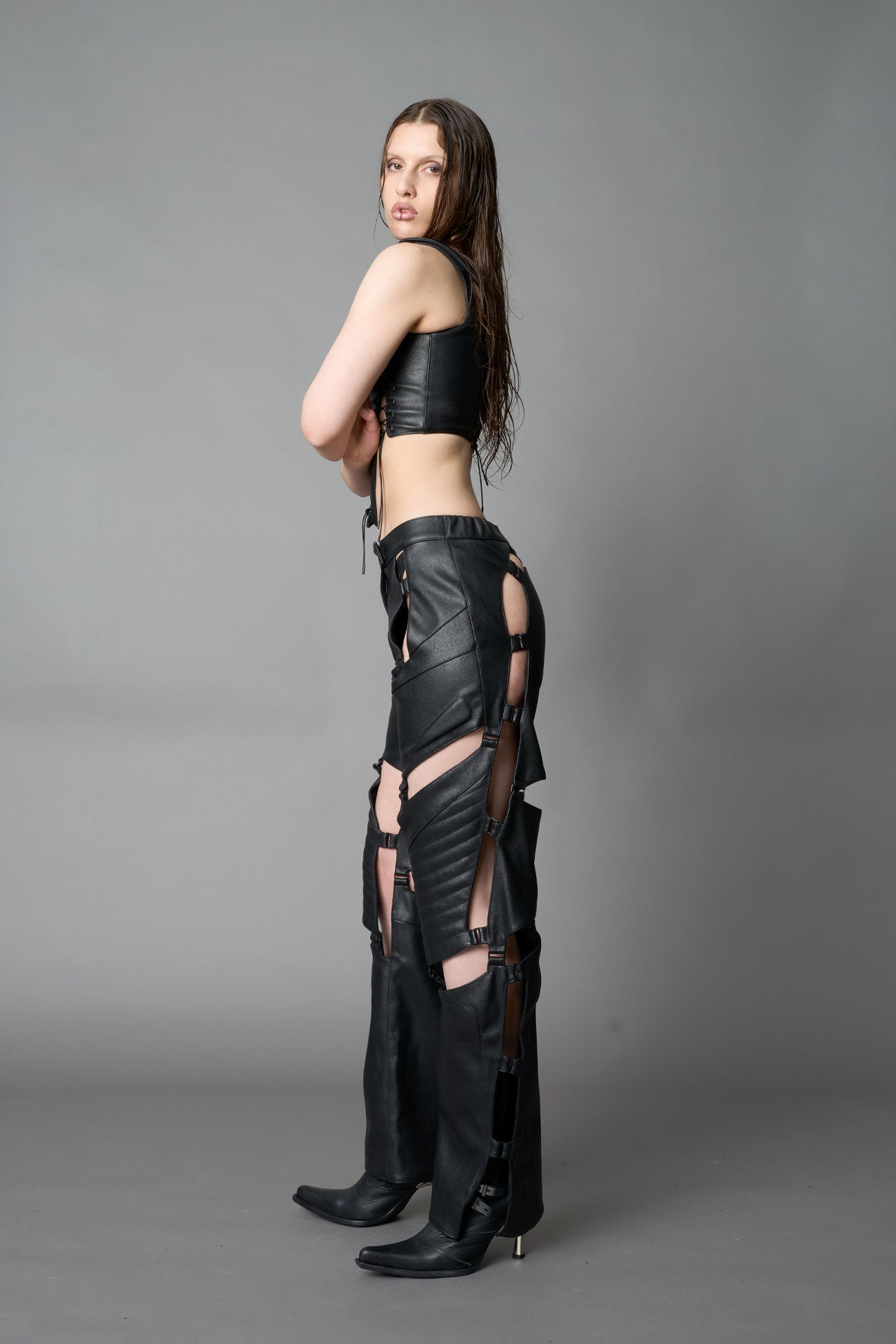 female Model wearing black vegan leather motocross-inspired pants with thigh cut-outs, strap harness details, quilted knee padding, and full-length leg gaiters; styled with a structured black bandeau top against a neutral background.