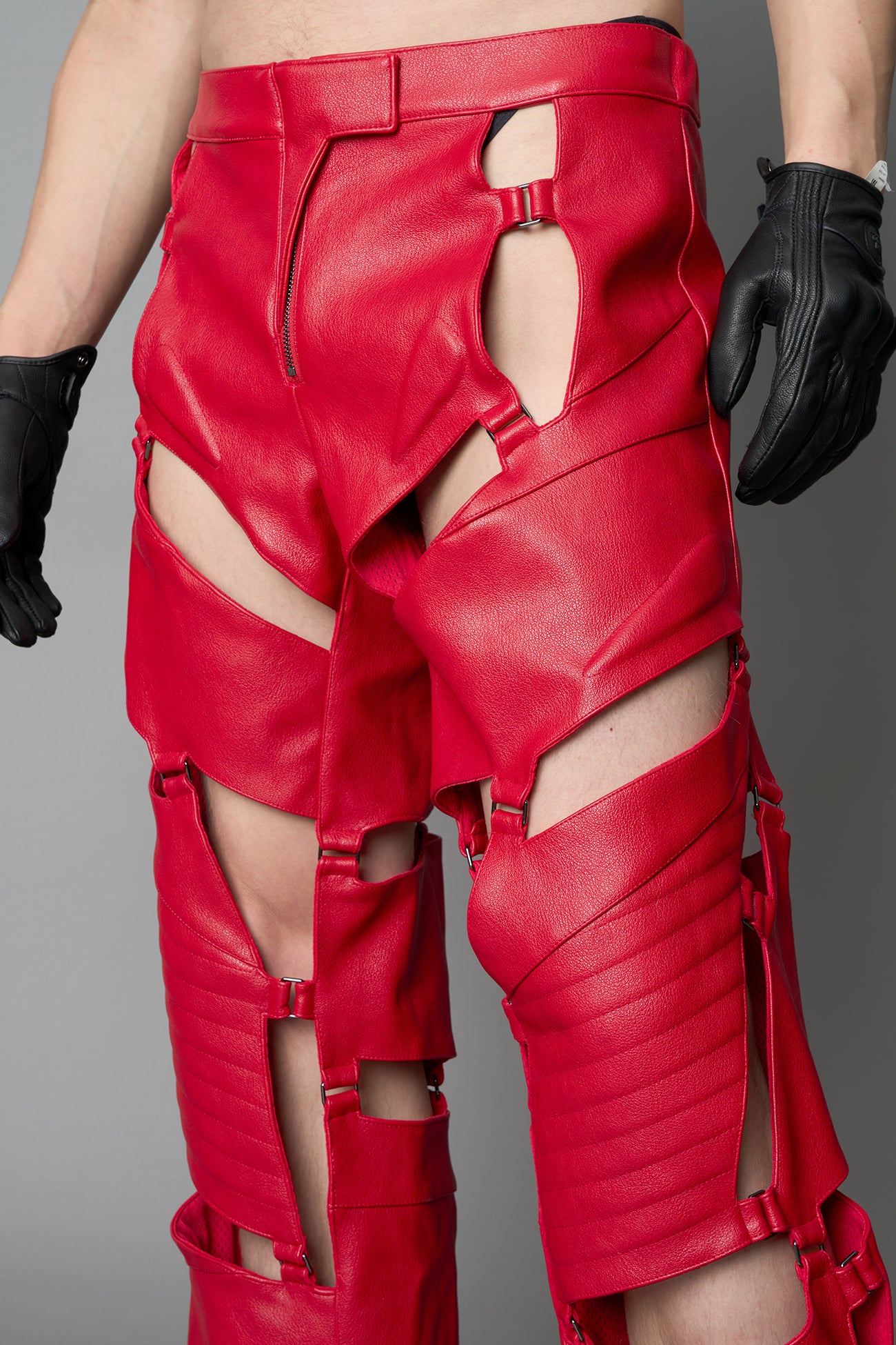 Model wearing red vegan leather motocross-inspired pants with thigh cut-outs, strap harness details, quilted knee padding, and full-length leg gaiters; styled with a structured black bandeau top against a neutral background.