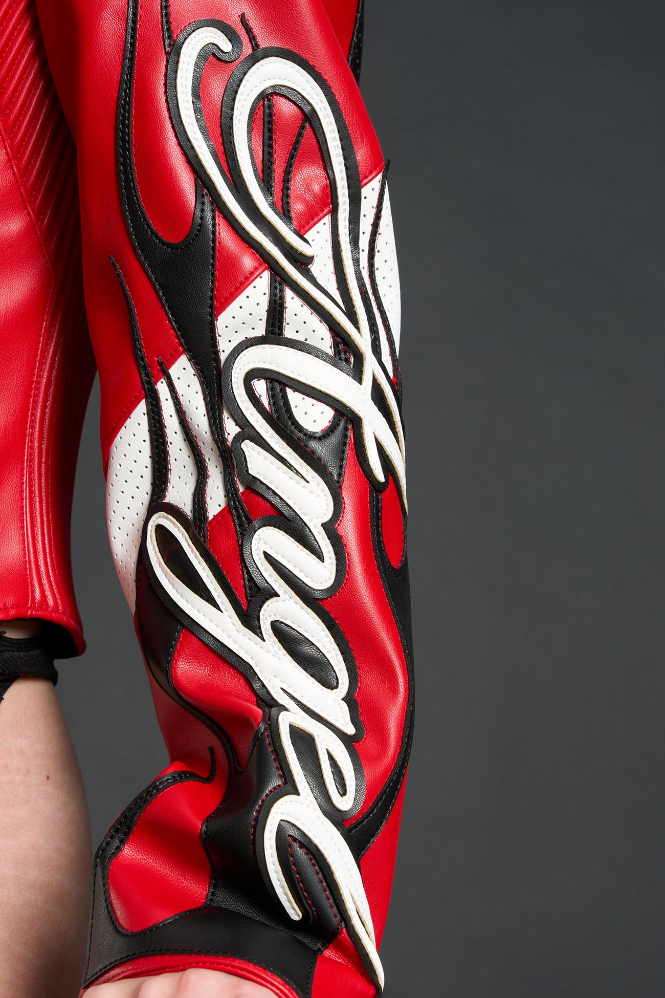 Model wears a motocross jacket-style vegan leather bodysuit with red motocross paneling, padded details, flame graphics, and ‘Hollywood Angel’ embroidery