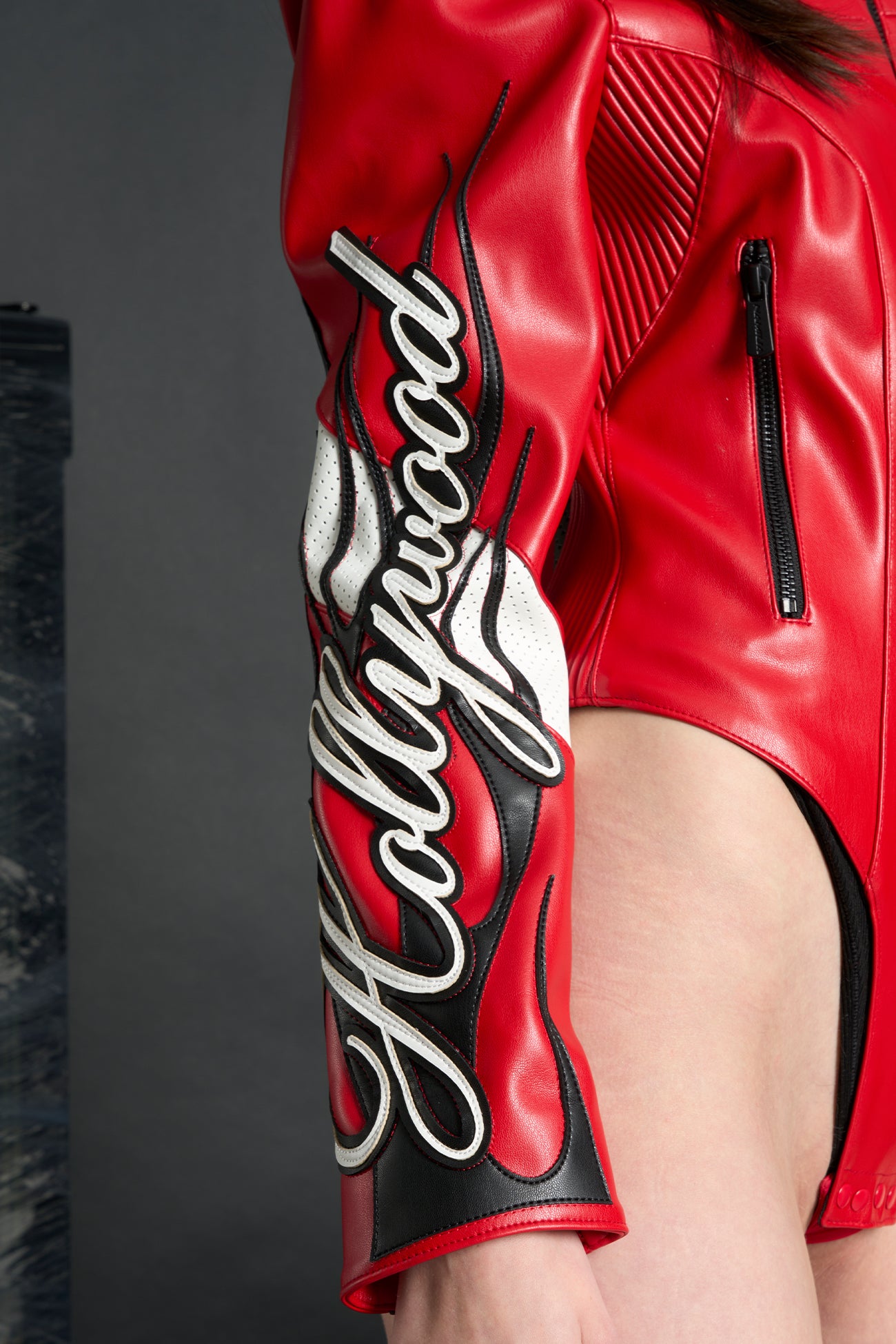 Model wears a motocross jacket-style vegan leather bodysuit with red motocross paneling, padded details, flame graphics, and ‘Hollywood Angel’ embroidery