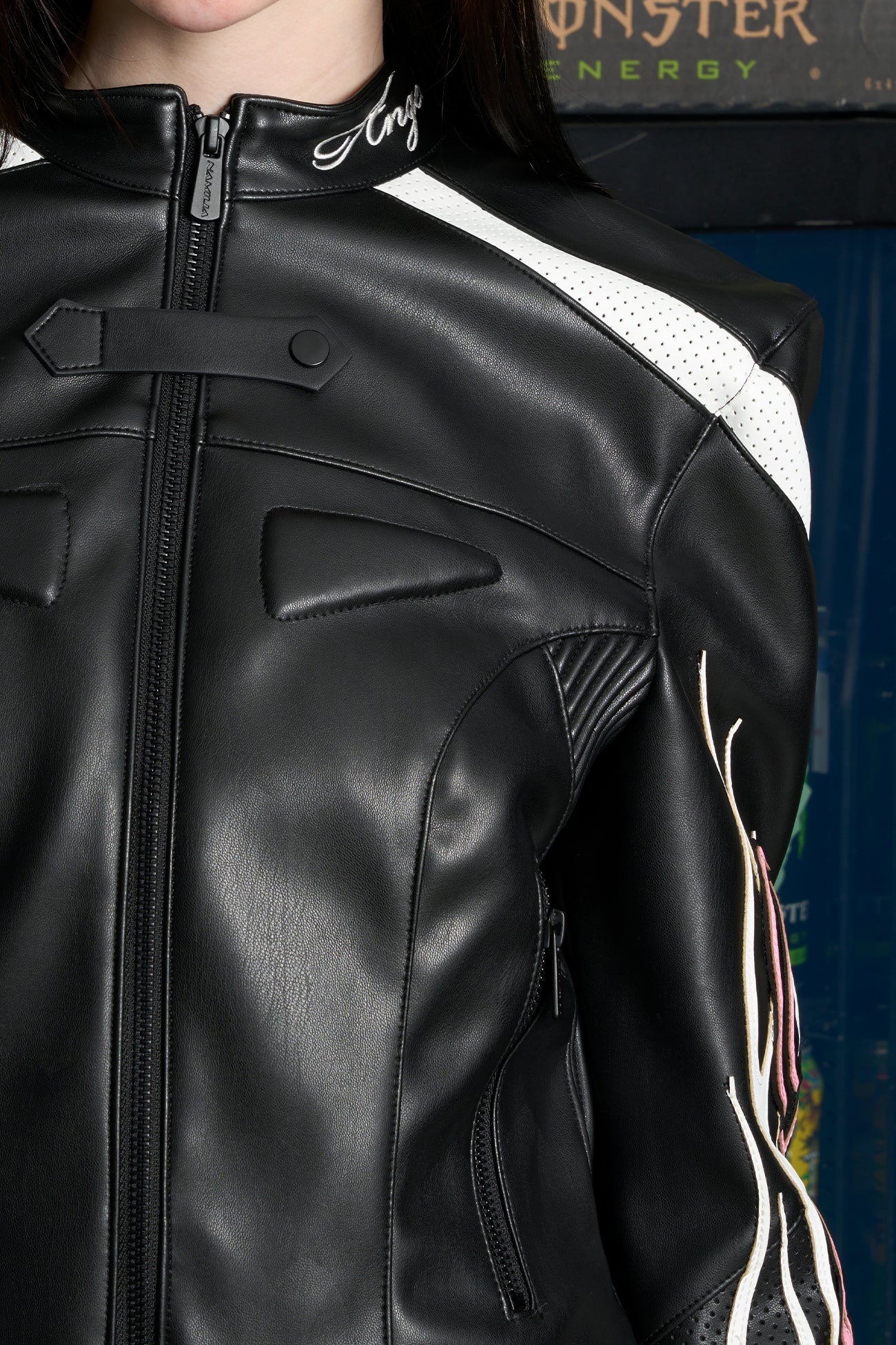Model wears a black motocross-style jacket made from vegan PU leather with flame topstitching, padded perforated paneling, topstiched sleeve slogan, ‘Angel’ embroidery on collar, and branded zippered front and pockets.