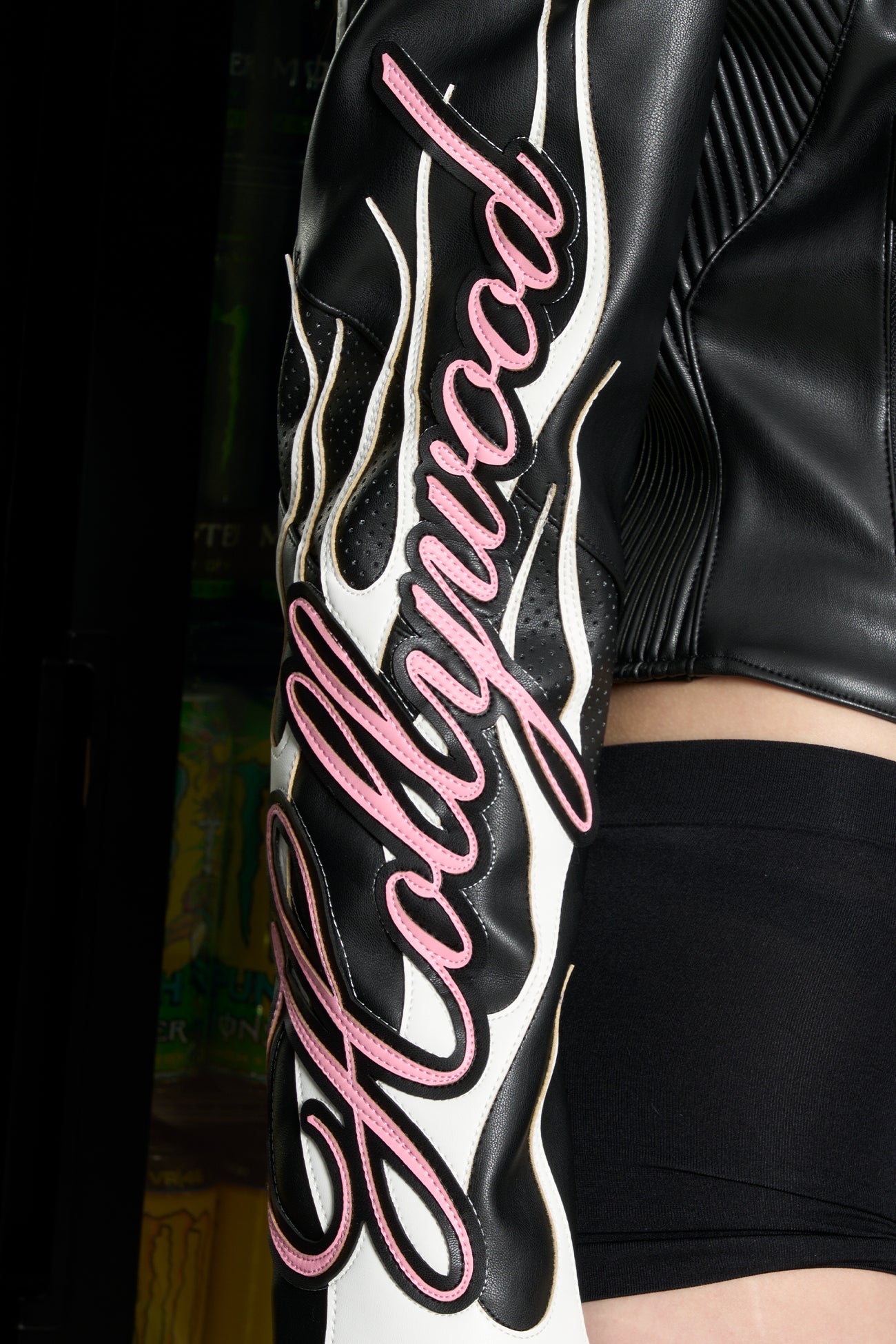 Model wears a black motocross-style jacket made from vegan PU leather with flame topstitching, padded perforated paneling, topstiched sleeve slogan, ‘Angel’ embroidery on collar, and branded zippered front and pockets.