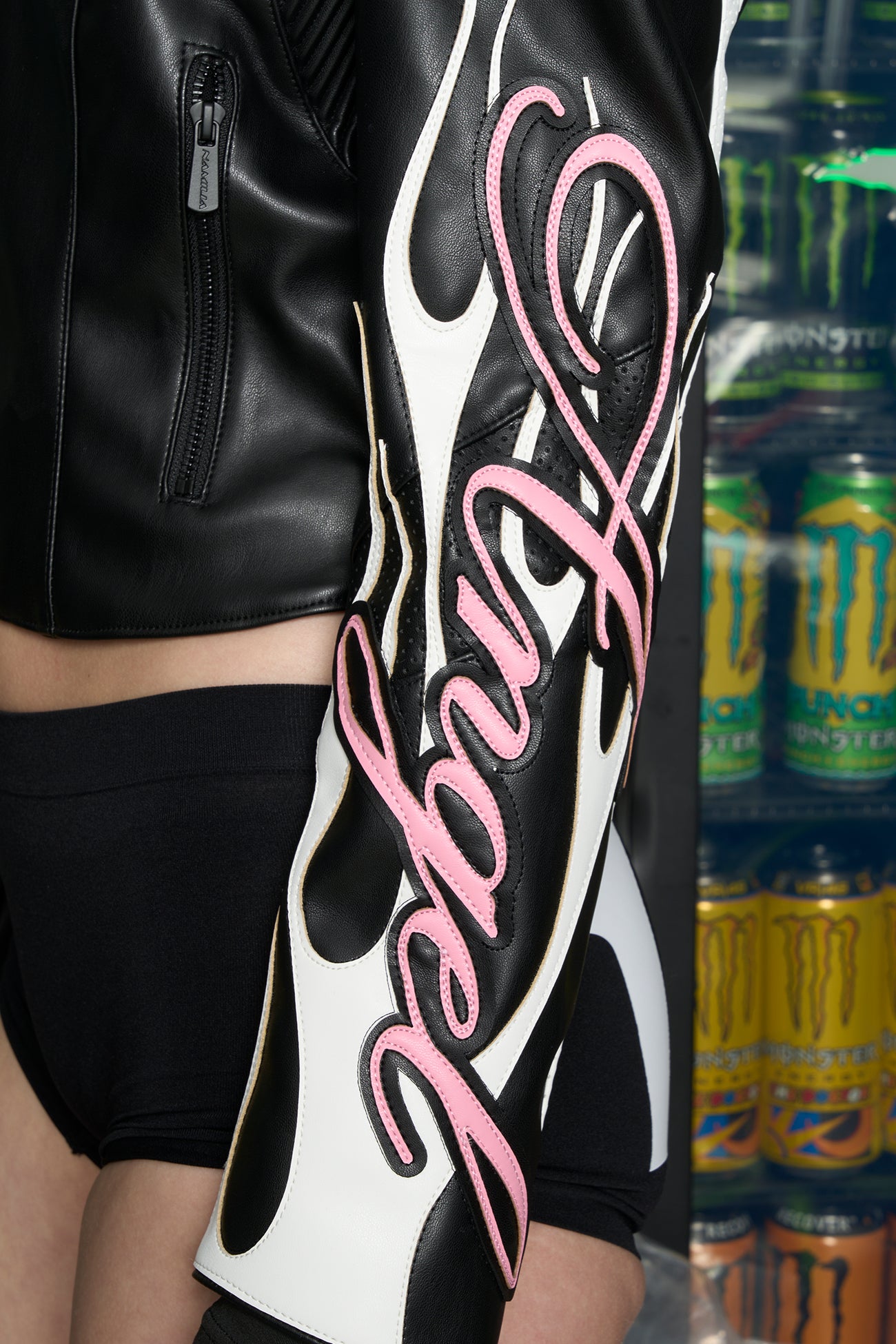 Model wears a black motocross-style jacket made from vegan PU leather with flame topstitching, padded perforated paneling, topstiched sleeve slogan, ‘Angel’ embroidery on collar, and branded zippered front and pockets.