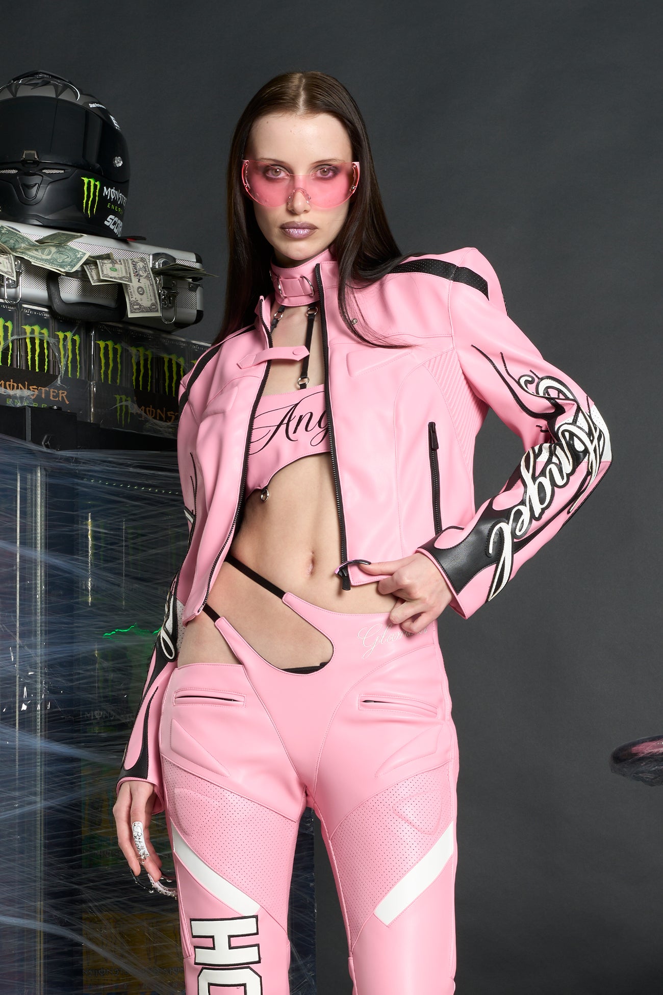 Model wears a pink motocross-style jacket made from vegan PU leather with flame topstitching, padded perforated paneling, topstiched sleeve slogan, ‘Angel’ embroidery on collar, and branded zippered front and pockets.