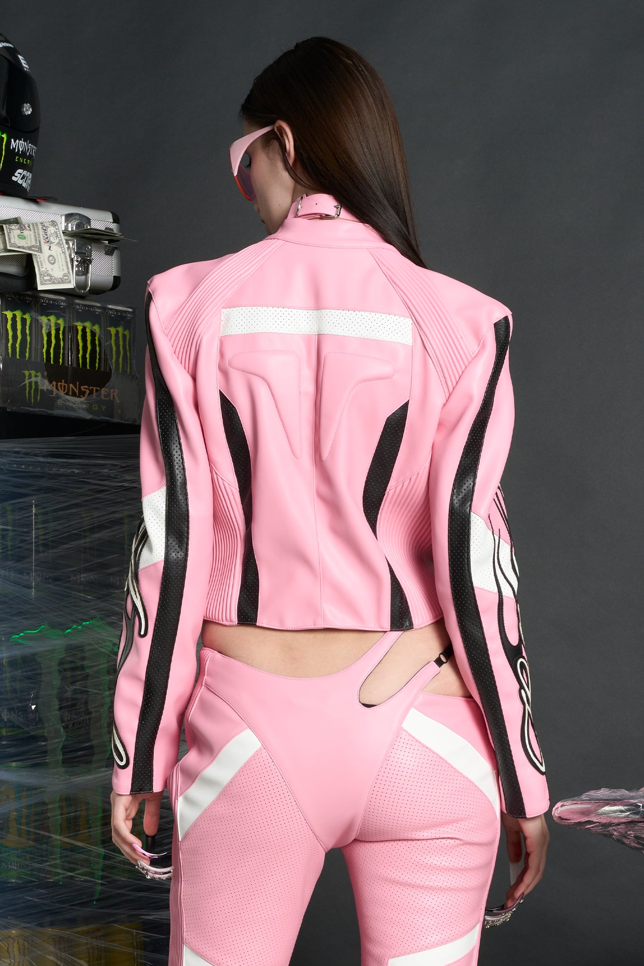 Model wears a pink motocross-style jacket made from vegan PU leather with flame topstitching, padded perforated paneling, topstiched sleeve slogan, ‘Angel’ embroidery on collar, and branded zippered front and pockets.